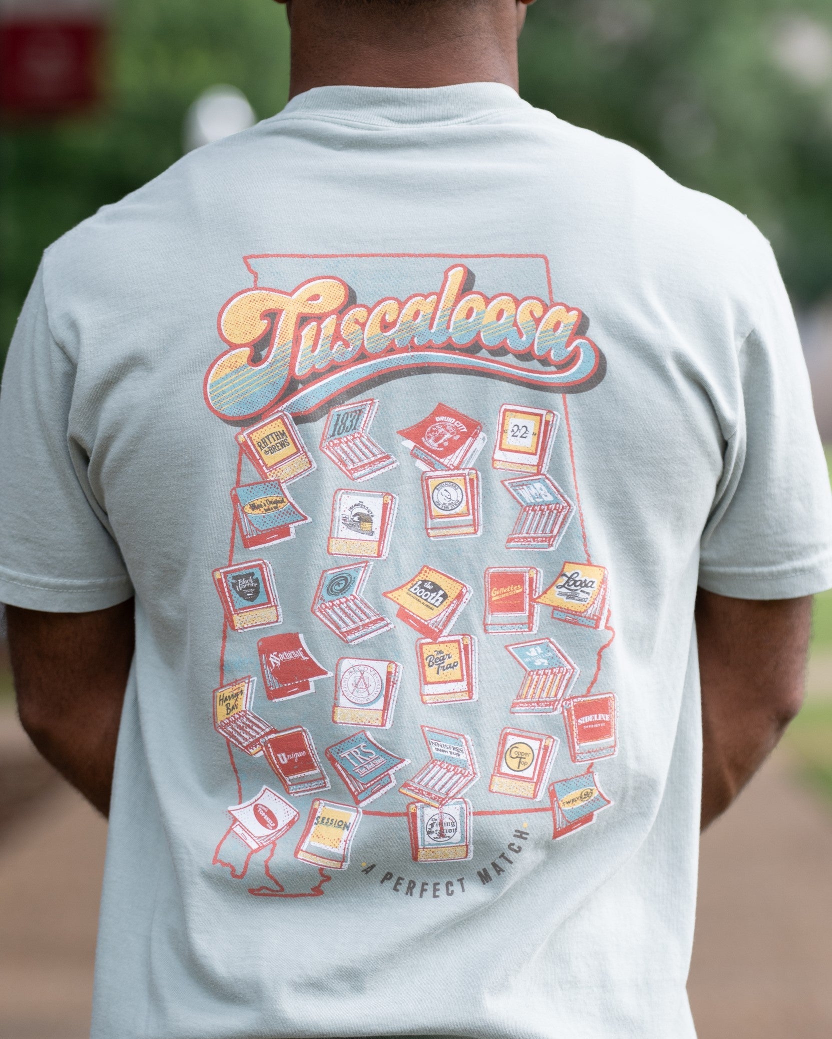 A Perfect Match Green Comfort Colors T-Shirt  with the text "Tuscaloosa" and images of matchbooks inside of an outline of the state of Alabama and smaller text reading "A PERFECT MATCH" outside the outline