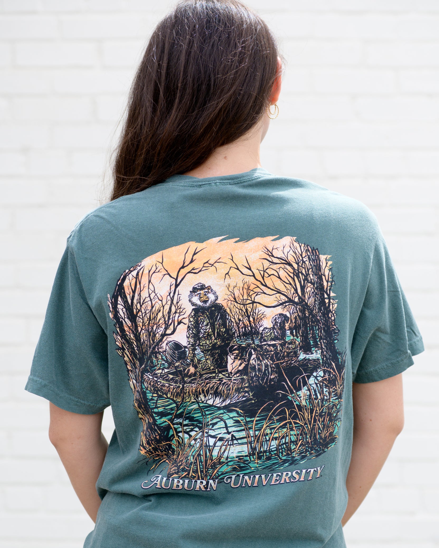 Auburn University Aubie Duck Hunt Blue Spruce Comfort Colors T-Shirt with an image of Aubie the Tiger wearing hunting gear with a dog in a boat in the swamp
