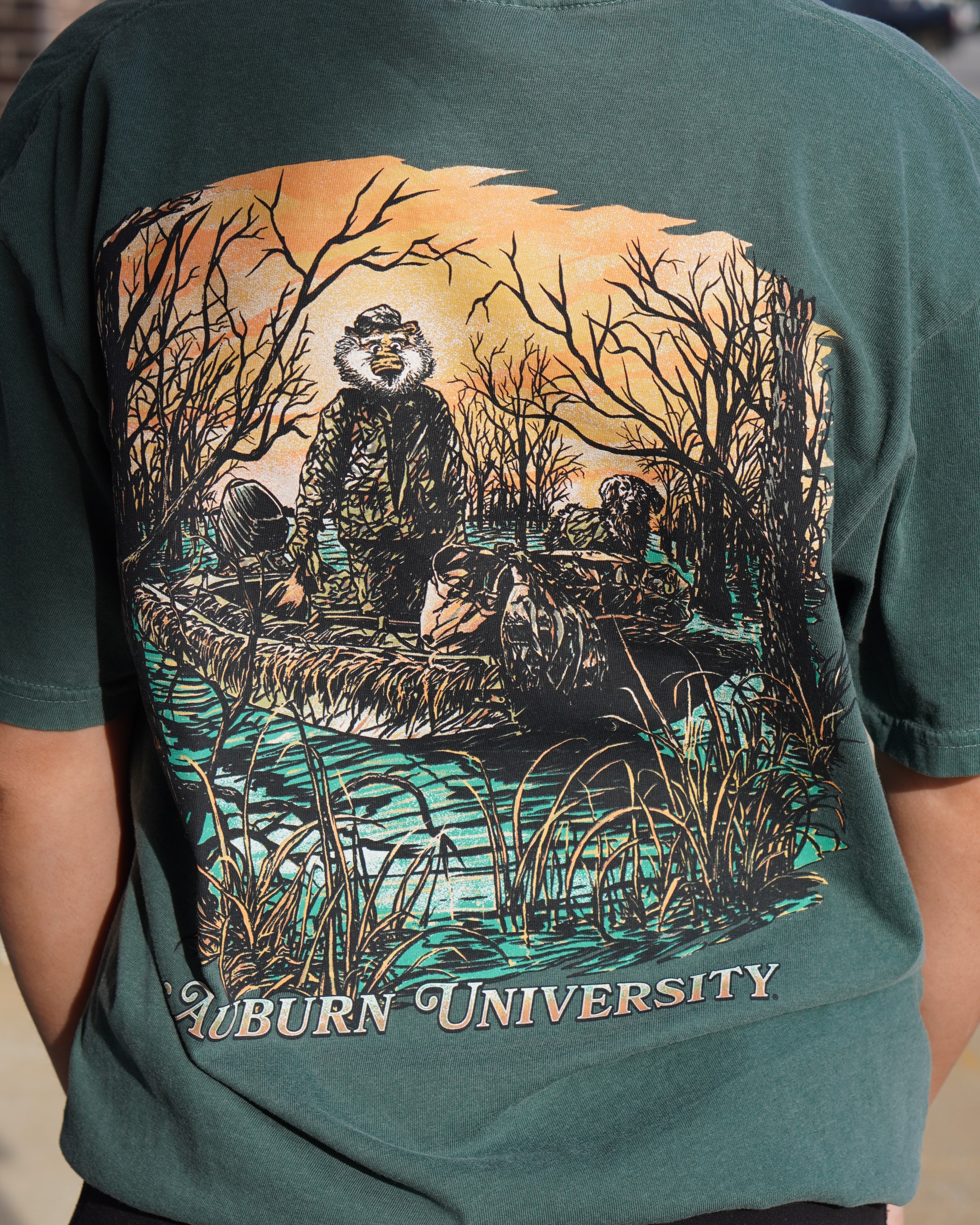 Auburn University Aubie Duck Hunt Blue Spruce Comfort Colors T-Shirt with an image of Aubie the Tiger wearing hunting gear with a dog in a boat in the swamp