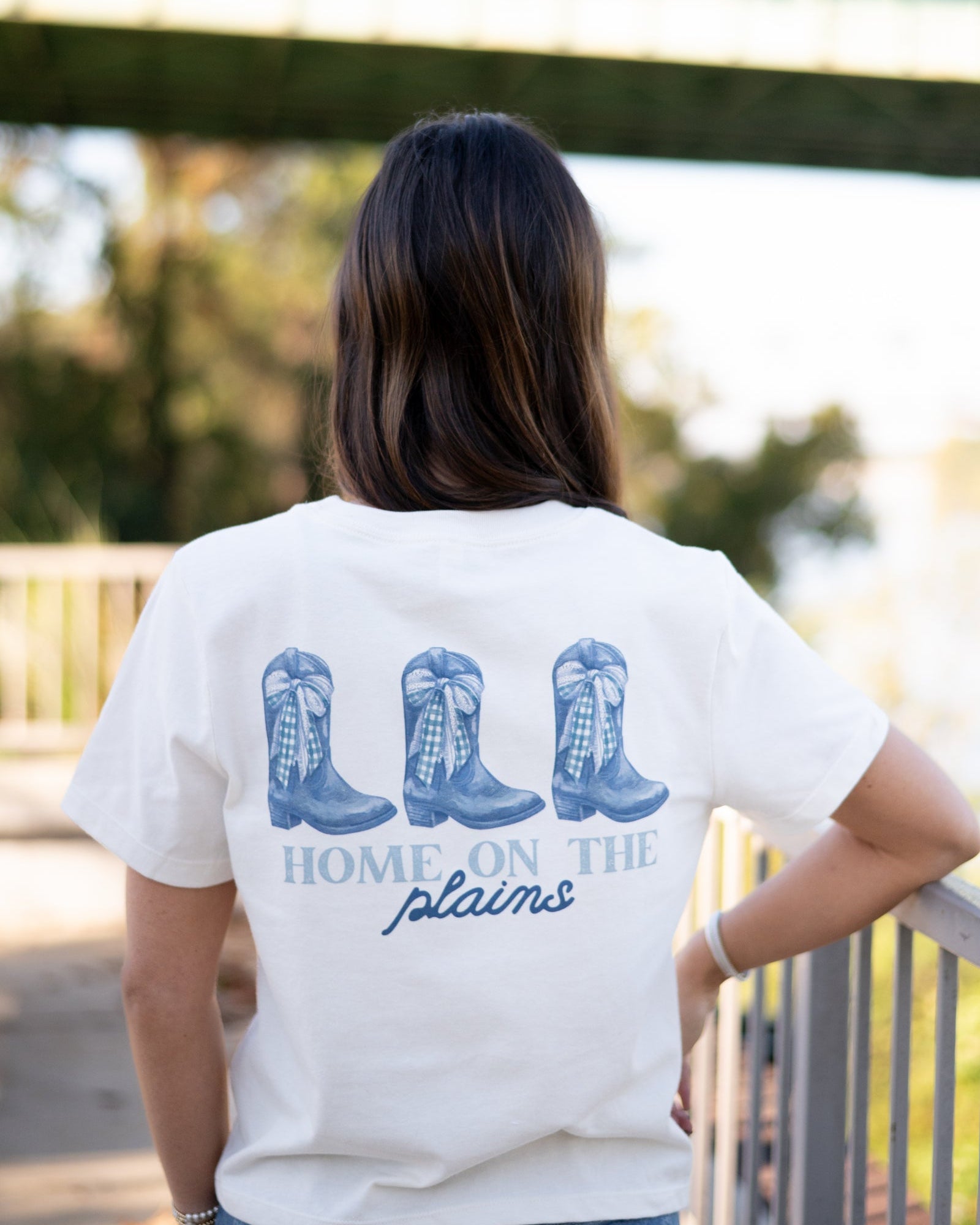 Auburn University White Ladies Tee with a vintage Blue illustration of cowgirl boots with bows tied on them above Blue text “Home On The Plains”