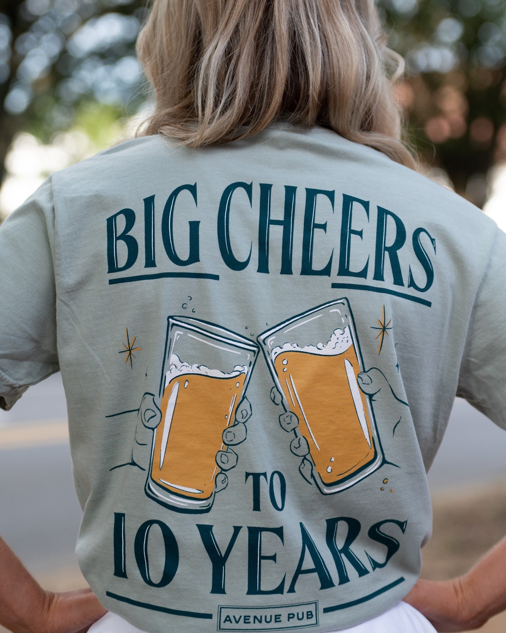 Avenue Pub's Beer Cheers Light Green Comfort Colors T-Shirt with an image of people clinking glasses of beer in between text "Big Cheers to 10 Years" (Back)