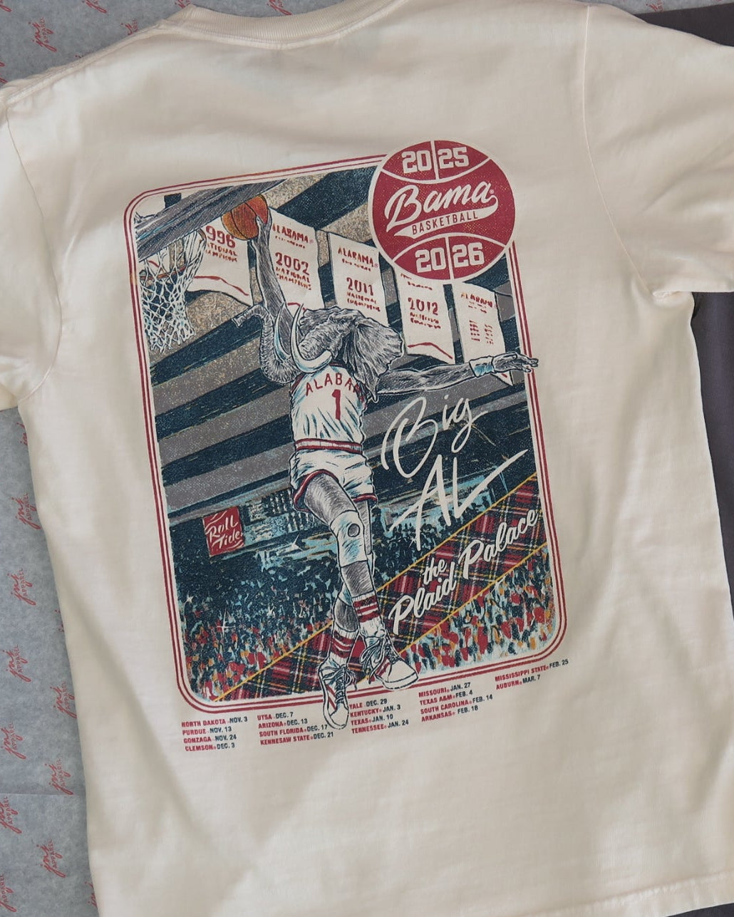 University of Alabama Ivory Shirt with an illustration of Big Al dunking a basketball next to text “Big Al” and “the Plaid Palace” above the 2025 Basketball Schedule