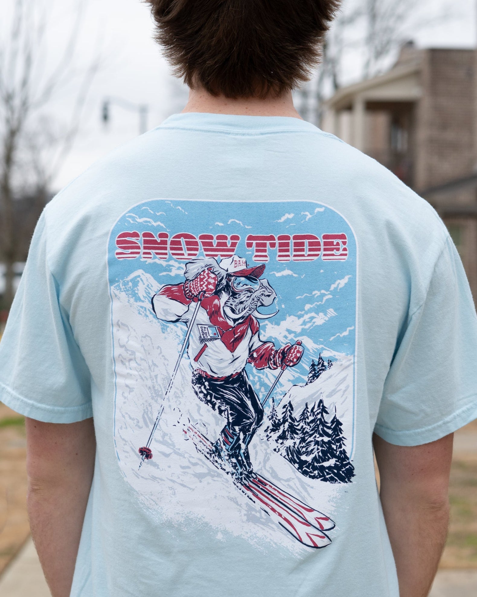 University of Alabama Light Blue T-Shirt with text "Snow Tide" above an image of Big Al in a ski outfit skiing down a snow-covered mountain