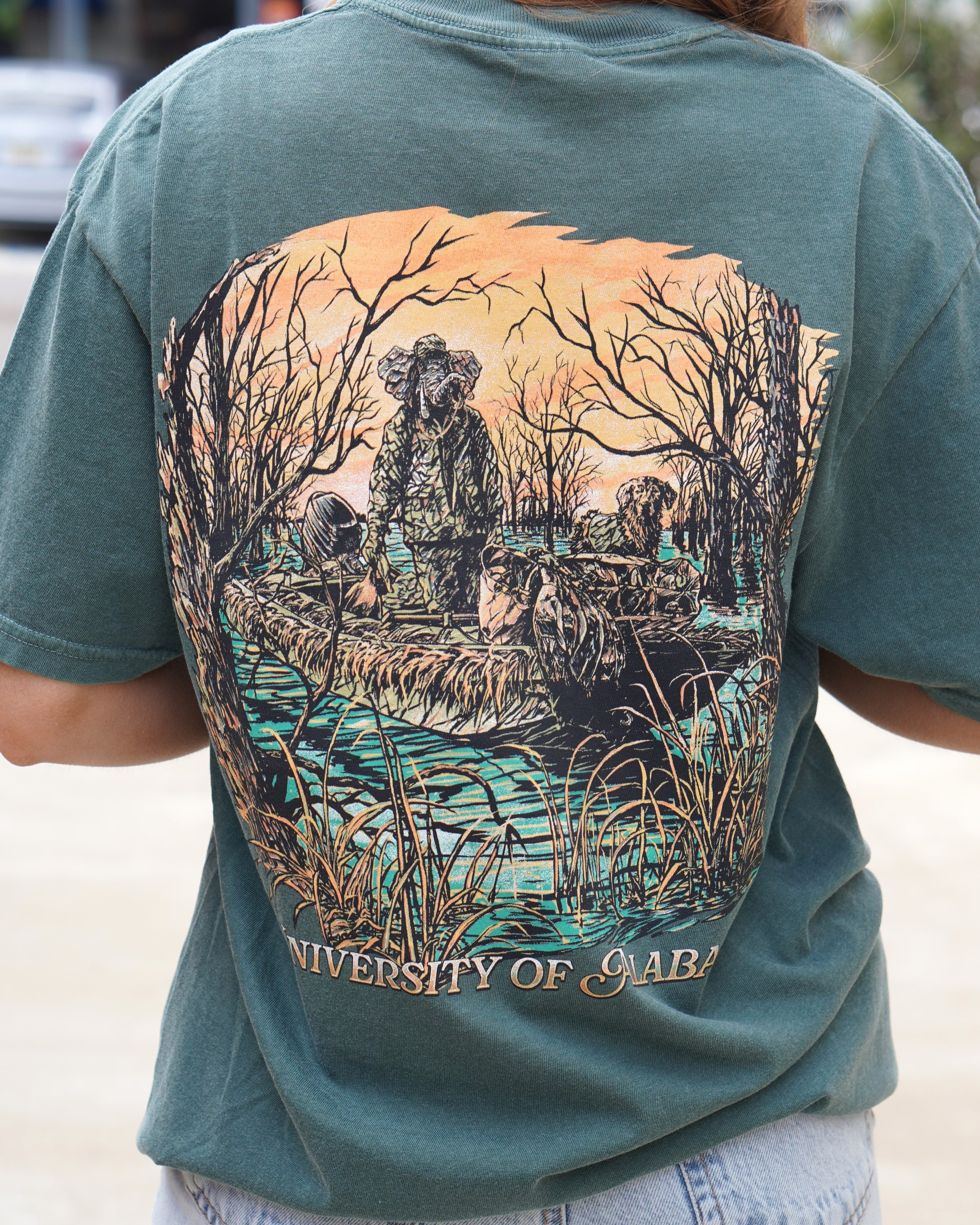 University of Alabama Big Al Duck Hunt Blue Spruce Comfort Colors T-Shirt with an image of Big Al wearing hunting gear with a dog in a boat in the swamp