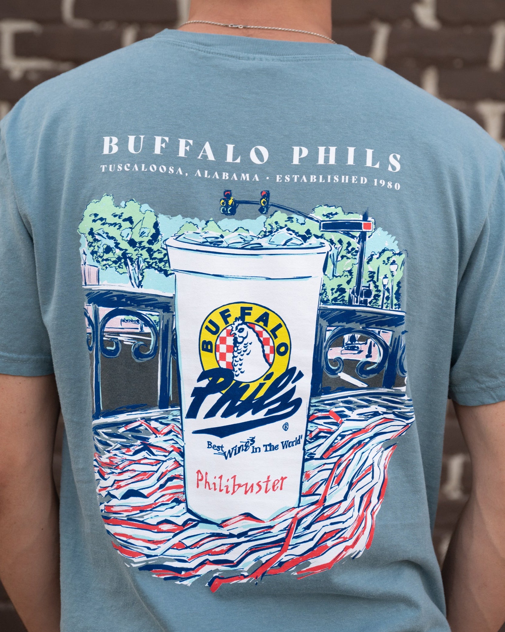 Ice Blue Buffalo Phil's T-Shirt with an image of a Buffalo Phil's cup sitting on top of a shaker on a table looking out on The Strip