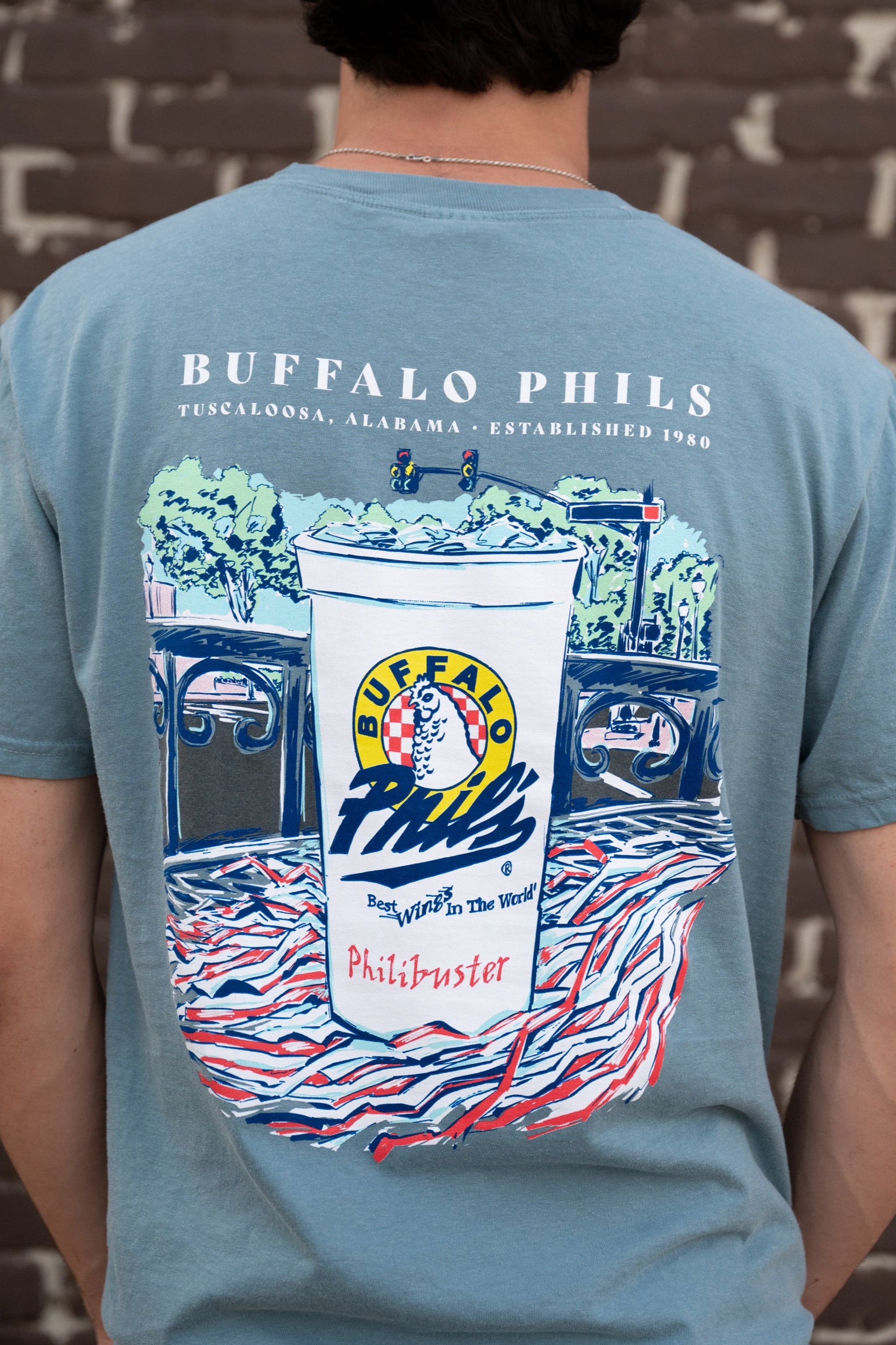 Ice Blue Buffalo Phil's T-Shirt with an image of a Buffalo Phil's cup sitting on top of a shaker on a table looking out on The Strip