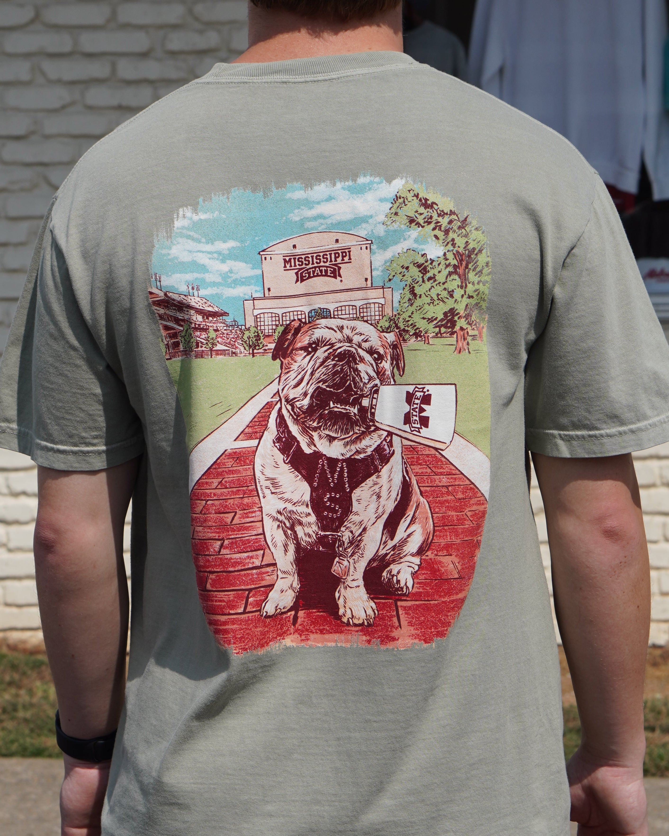 Mississippi State Tan T-Shirt with an image of Bully the bulldog holding a cowbell in it’s mouth on the brick walk into campus