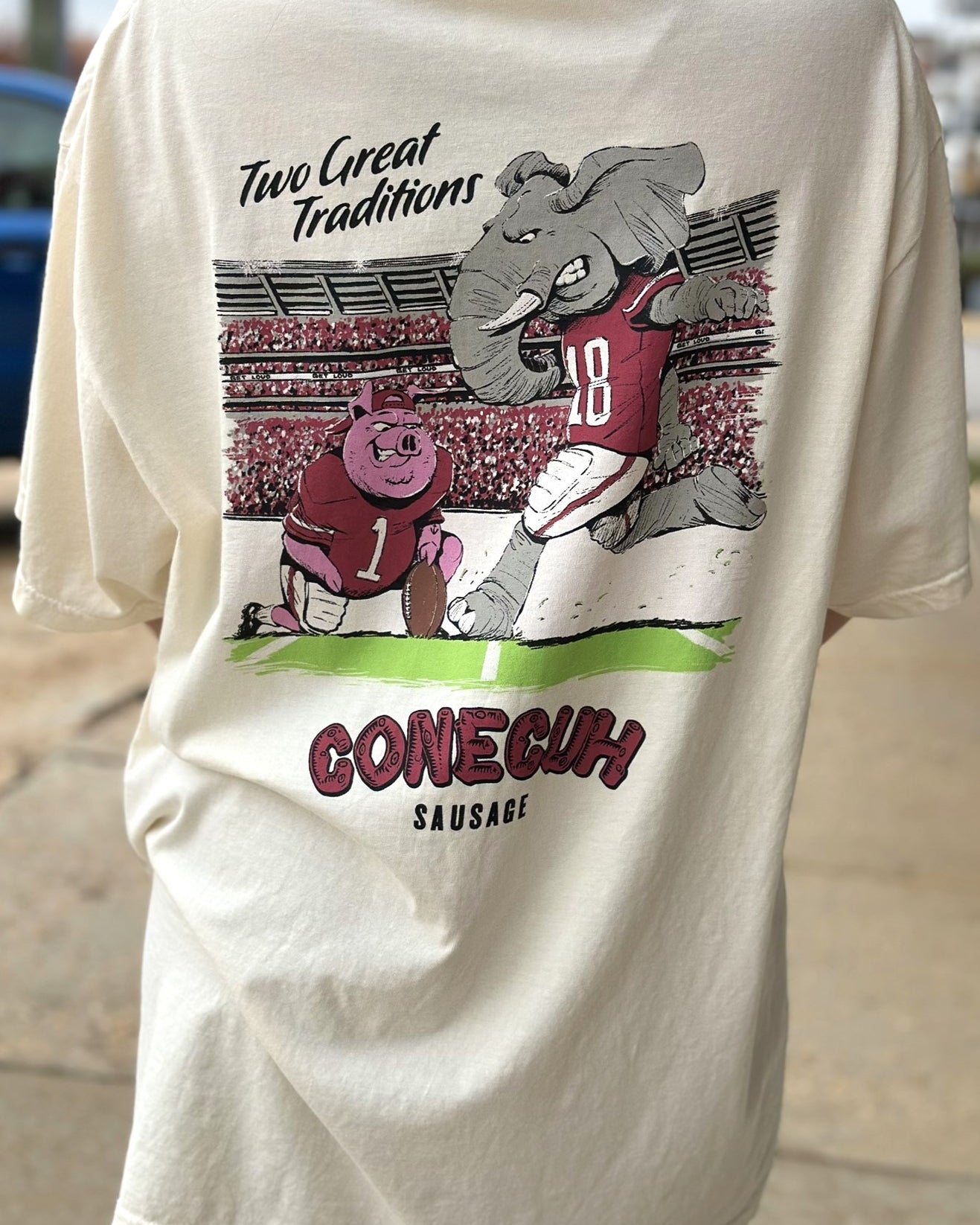 University of Alabama Conecuh Traditions Ivory Comfort Colors T-Shirt with Black text "Two Great Traditions" above an illustration of a pig holding a football for Big Al to kick