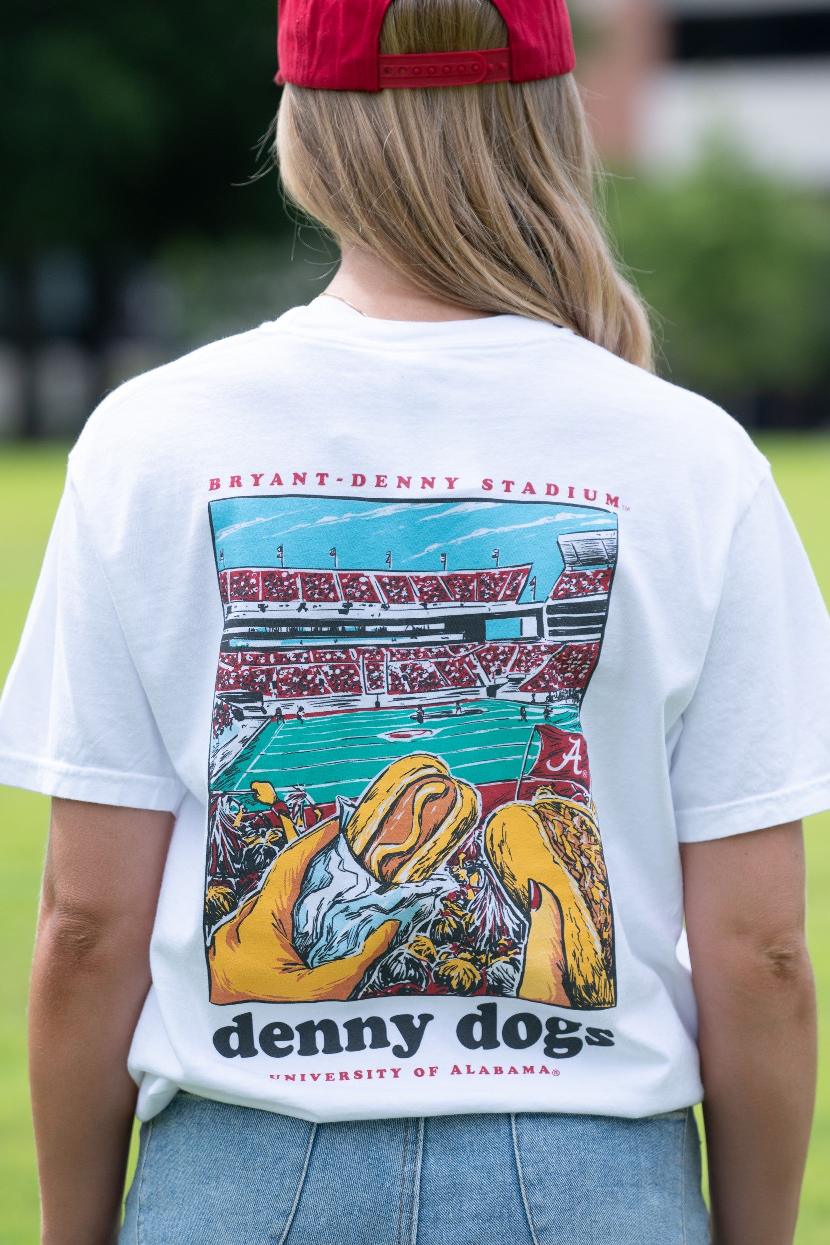 University of Alabama White Denny Dogs Comfort Colors T-Shirt with an image of two hands holding hot dogs at Bryant- Denny Stadium above the Black text "denny dogs"