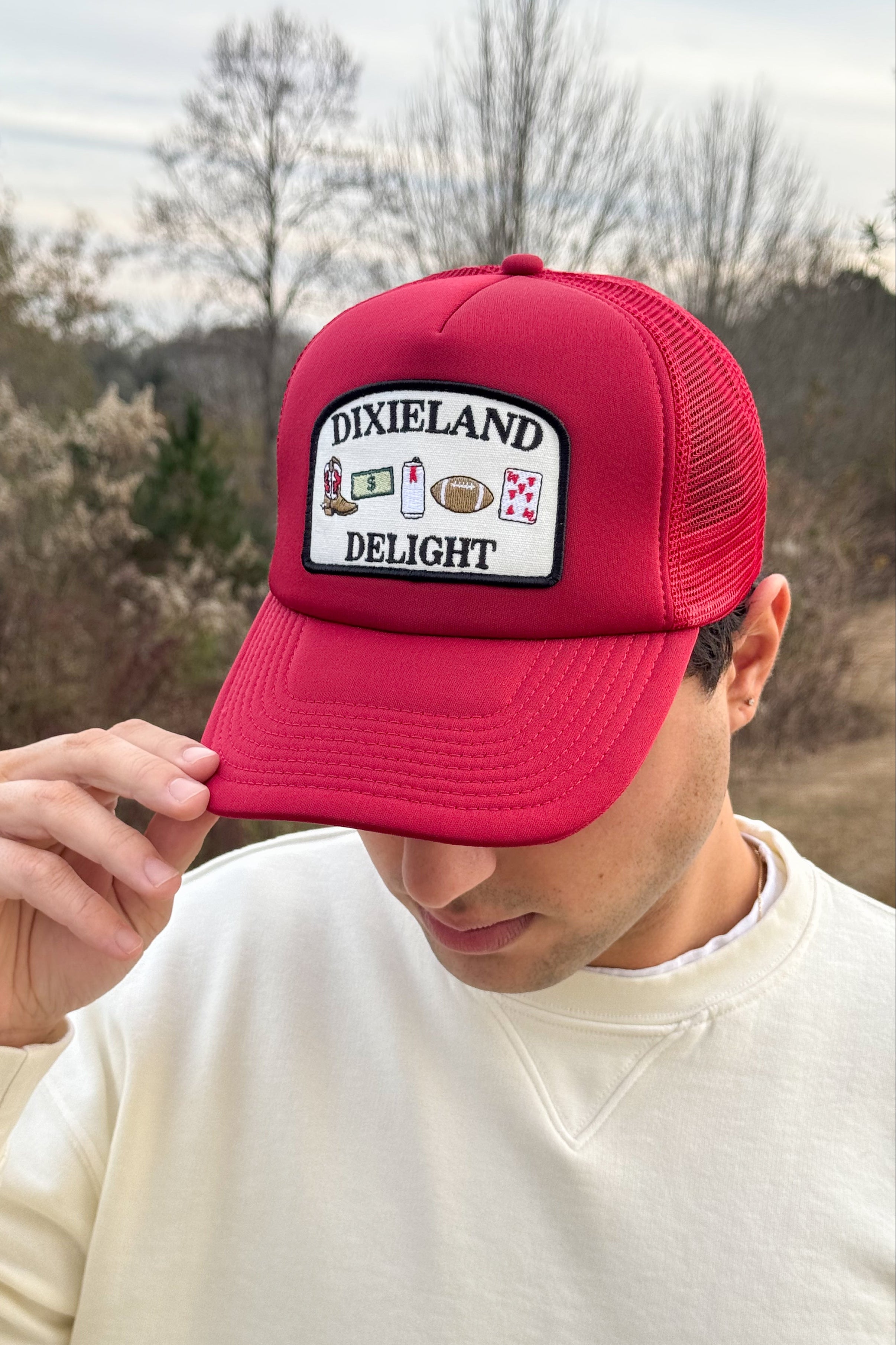 University of Alabama Red Trucker Hat with a patch featuring text “Dixieland” above and “Delight” below drawings of a cowboy boot, money, drink can, football, and playing card