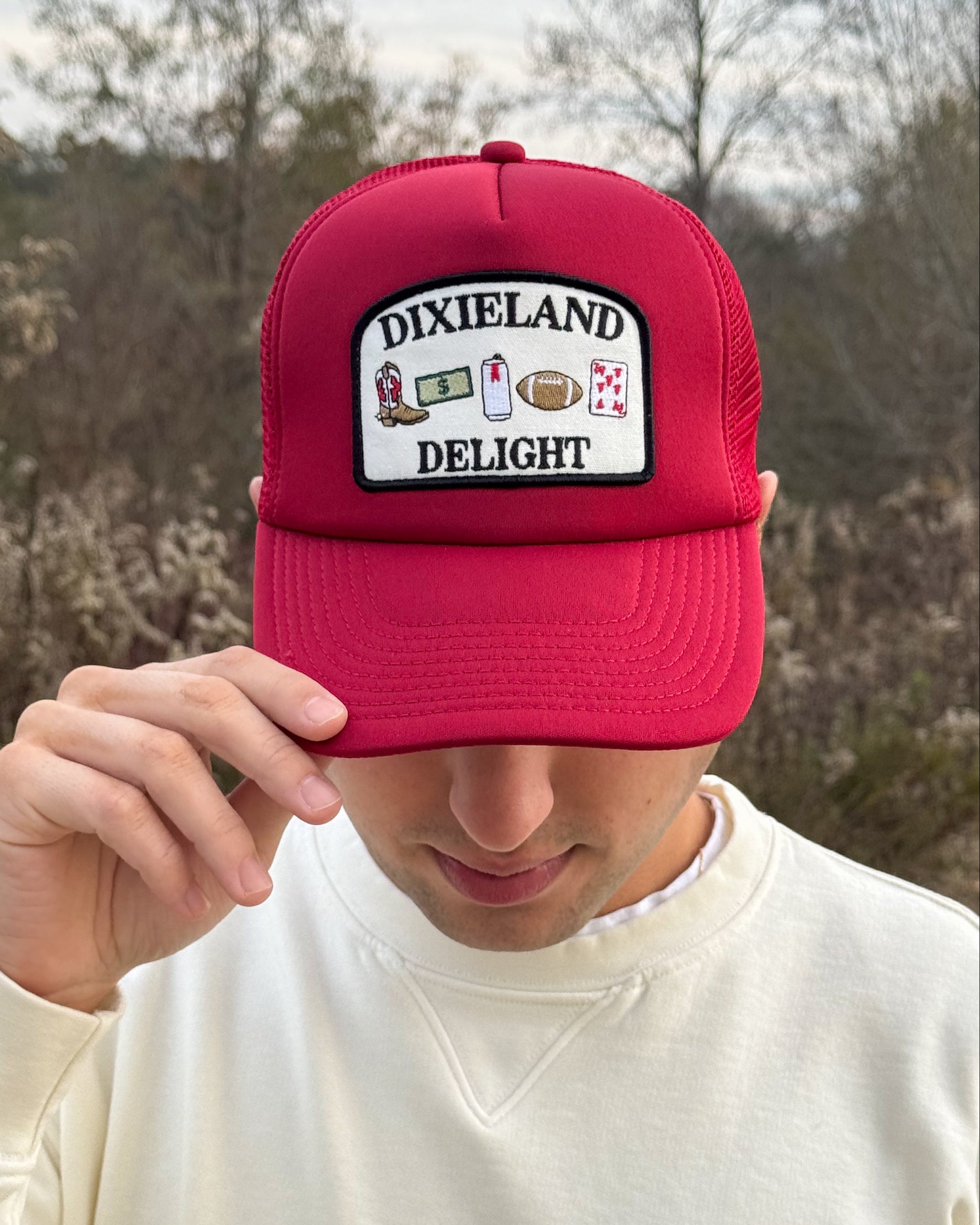 University of Alabama Red Trucker Hat with a patch featuring text “Dixieland” above and “Delight” below drawings of a cowboy boot, money, drink can, football, and playing card