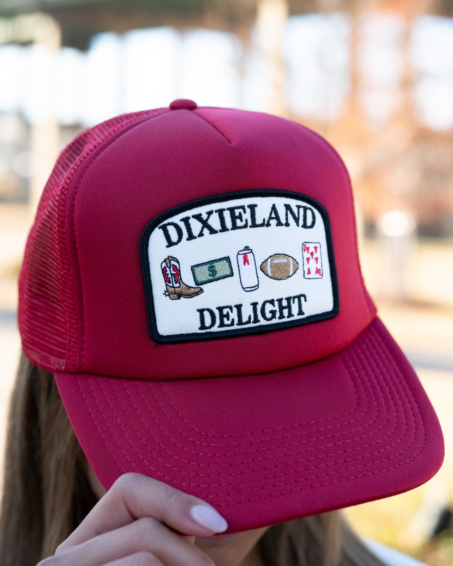 University of Alabama Red Trucker Hat with a patch featuring text “Dixieland” above and “Delight” below drawings of a cowboy boot, money, drink can, football, and playing card