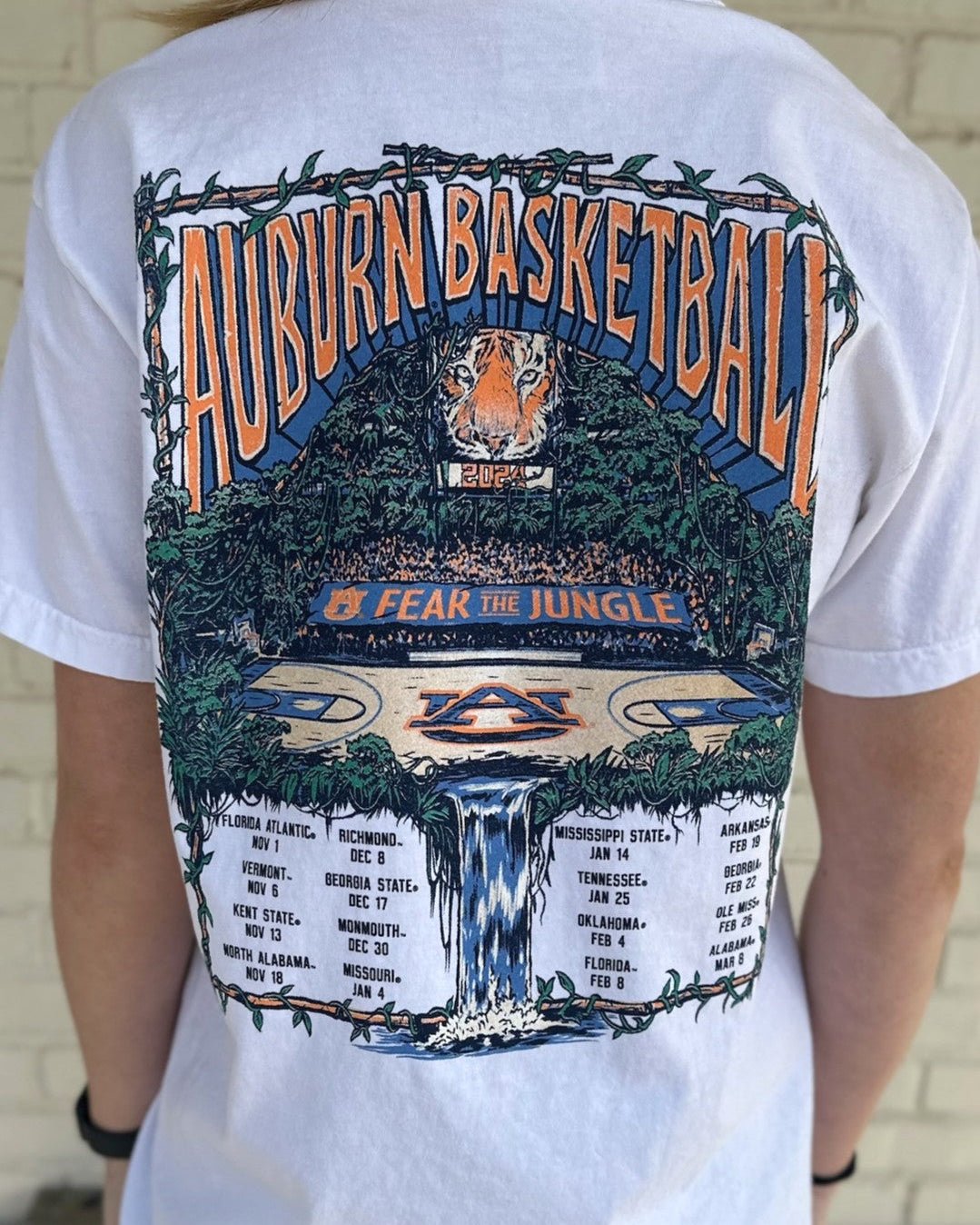 Auburn University White Fear The Jungle T-Shirt with Orange text "Auburn Basketball" and an image of the Auburn basketball court in the middle of a jungle with a waterfall spilling into the game schedule