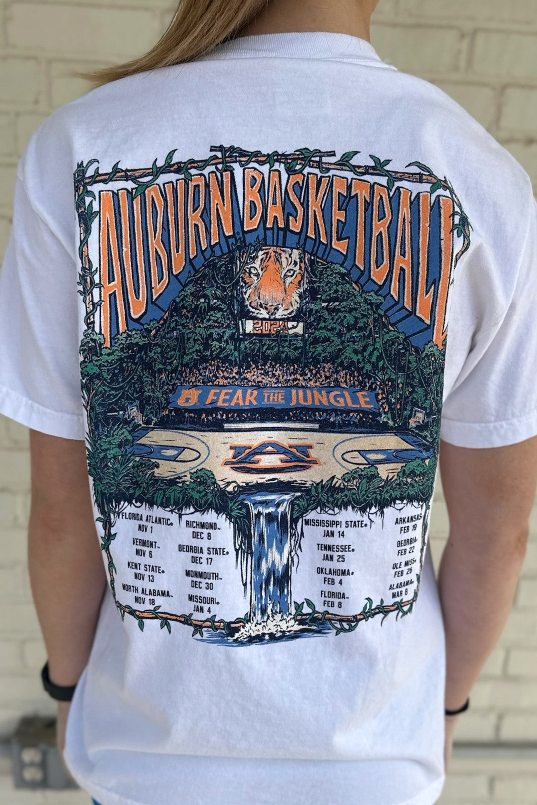 Auburn University White Fear The Jungle T-Shirt with Orange text "Auburn Basketball" and an image of the Auburn basketball court in the middle of a jungle with a waterfall spilling into the game schedule