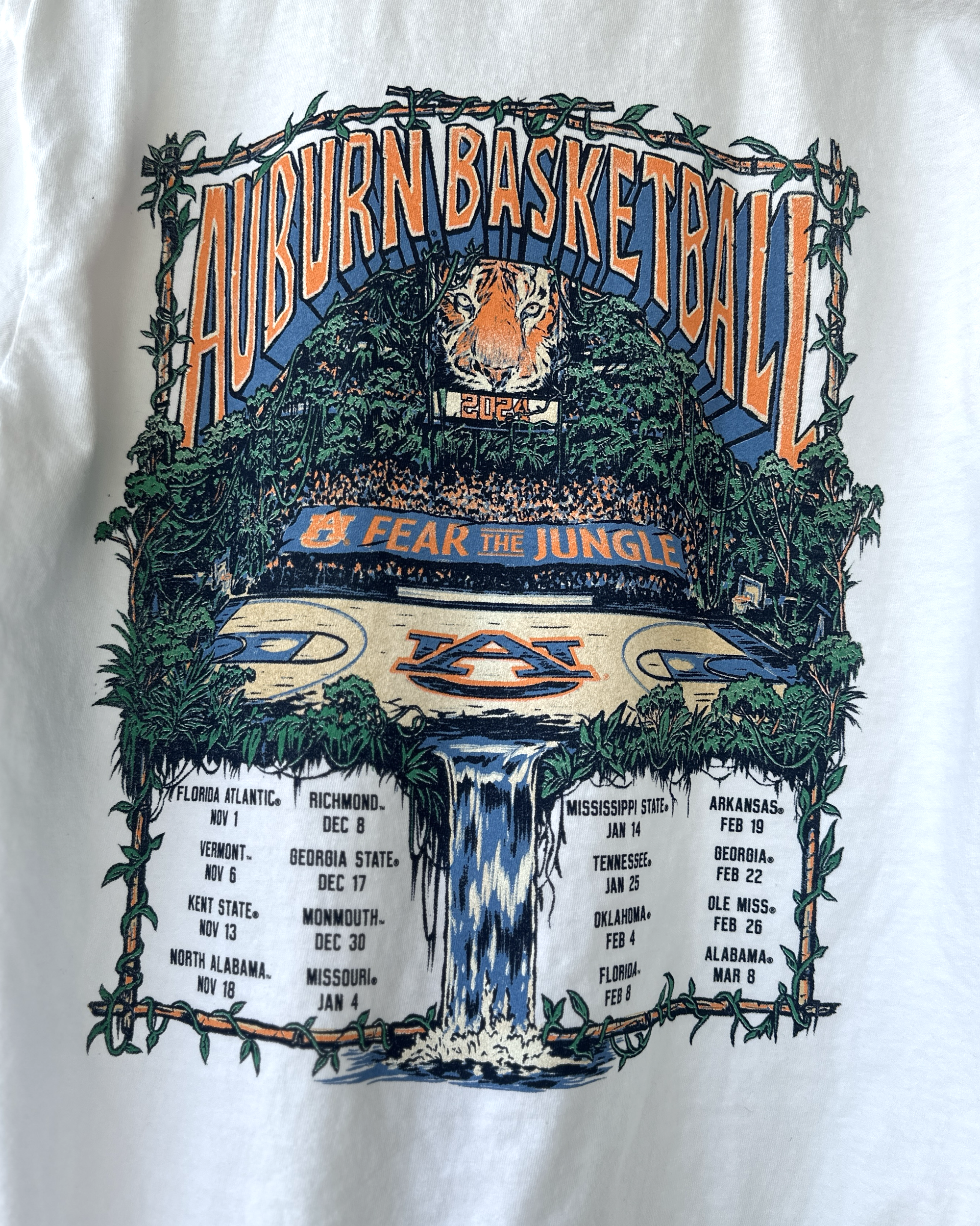 Auburn University White Fear The Jungle T-Shirt with Orange text "Auburn Basketball" and an image of the Auburn basketball court in the middle of a jungle with a waterfall spilling into the game schedule