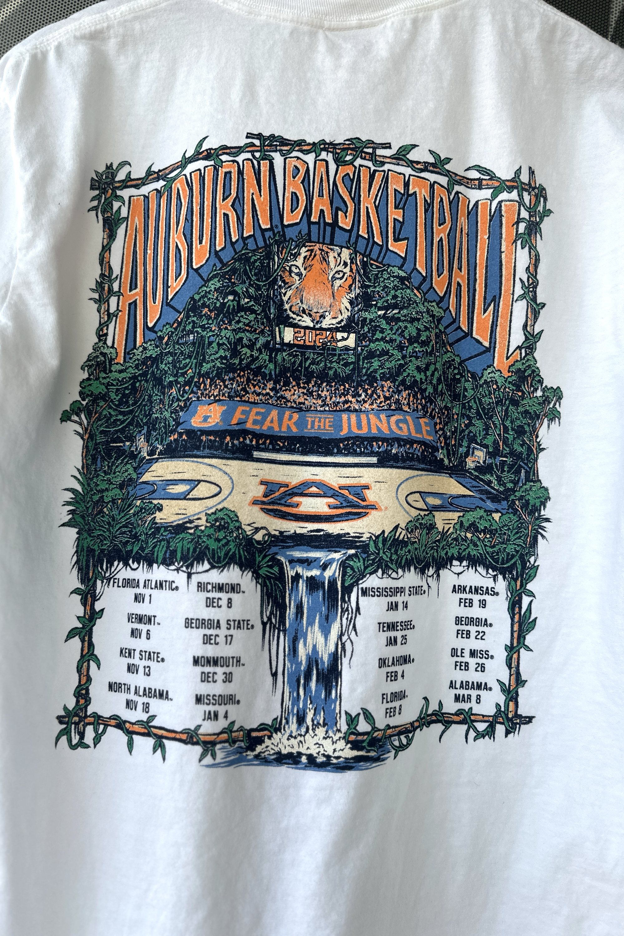 Auburn University White Fear The Jungle T-Shirt with Orange text "Auburn Basketball" and an image of the Auburn basketball court in the middle of a jungle with a waterfall spilling into the game schedule