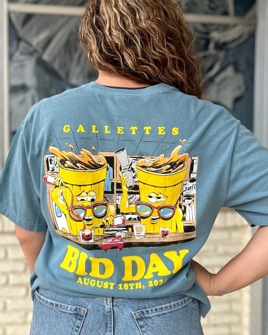 Gallettes Blue Bid Day T-Shirt with two yellow cups wearing sunglasses sitting at a bar and pouring themselves drinks above Yellow text "Bid Day August 18th, 2024"