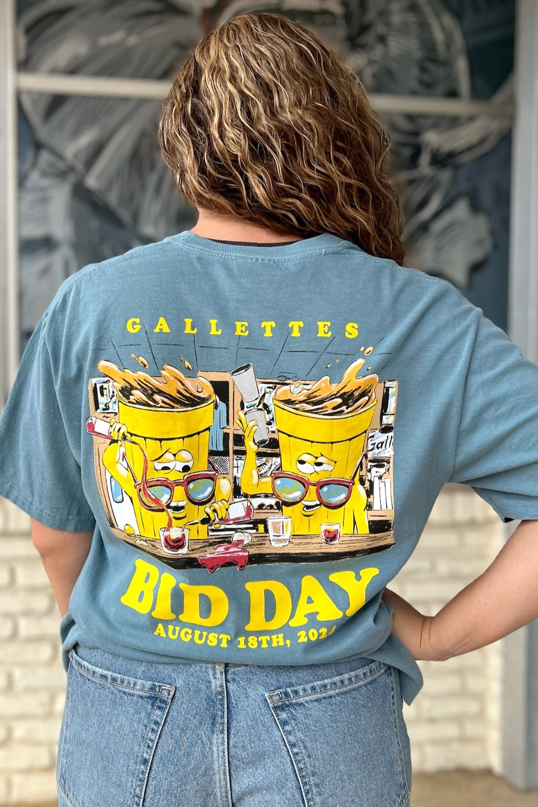 Gallettes Blue Bid Day T-Shirt with two yellow cups wearing sunglasses sitting at a bar and pouring themselves drinks above Yellow text "Bid Day August 18th, 2024"