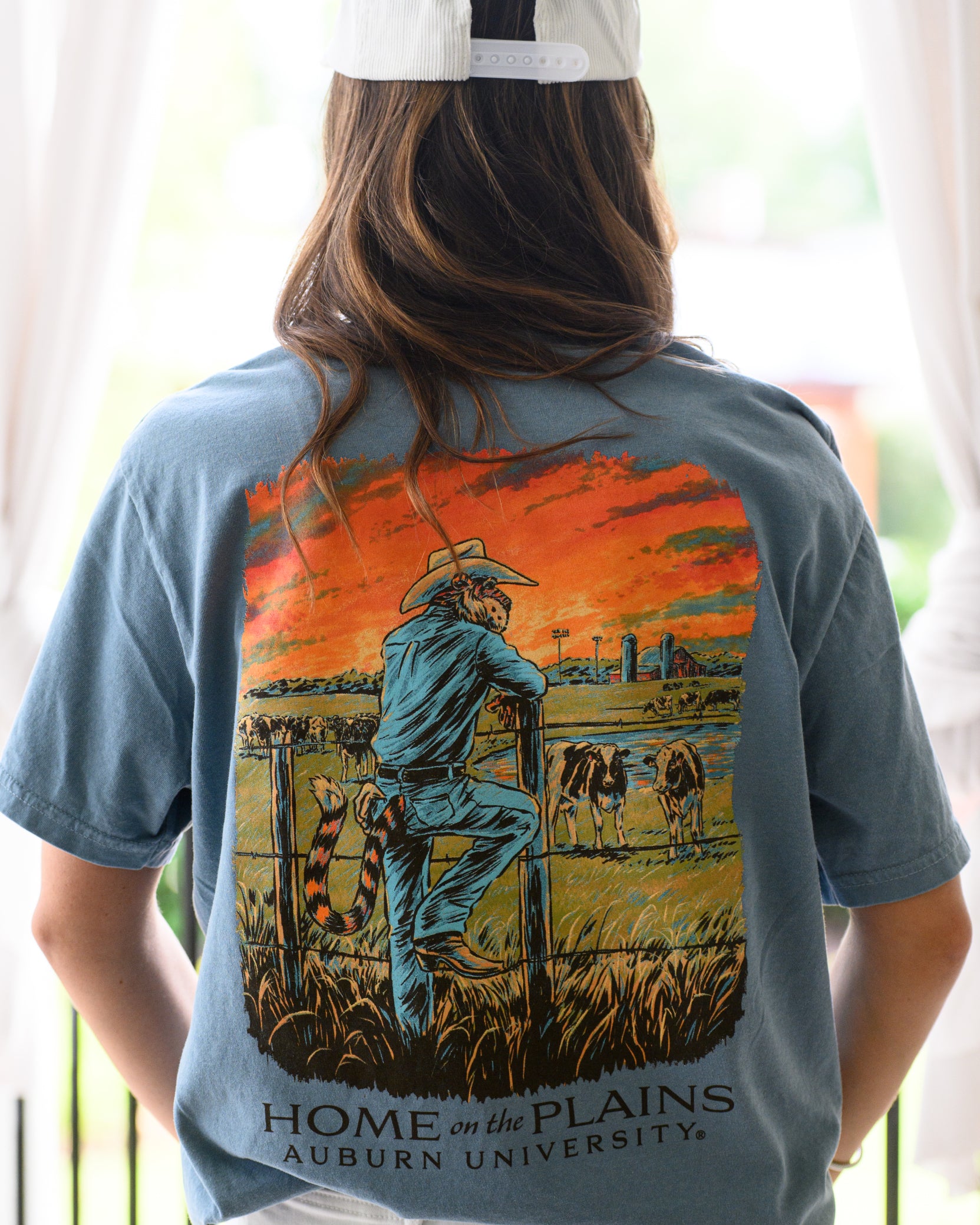 Auburn University Blue T-Shirt with an image of Aubie wearing cowboy clothes and watching over cattle at sunrise above Black text “Home on the Plains”
