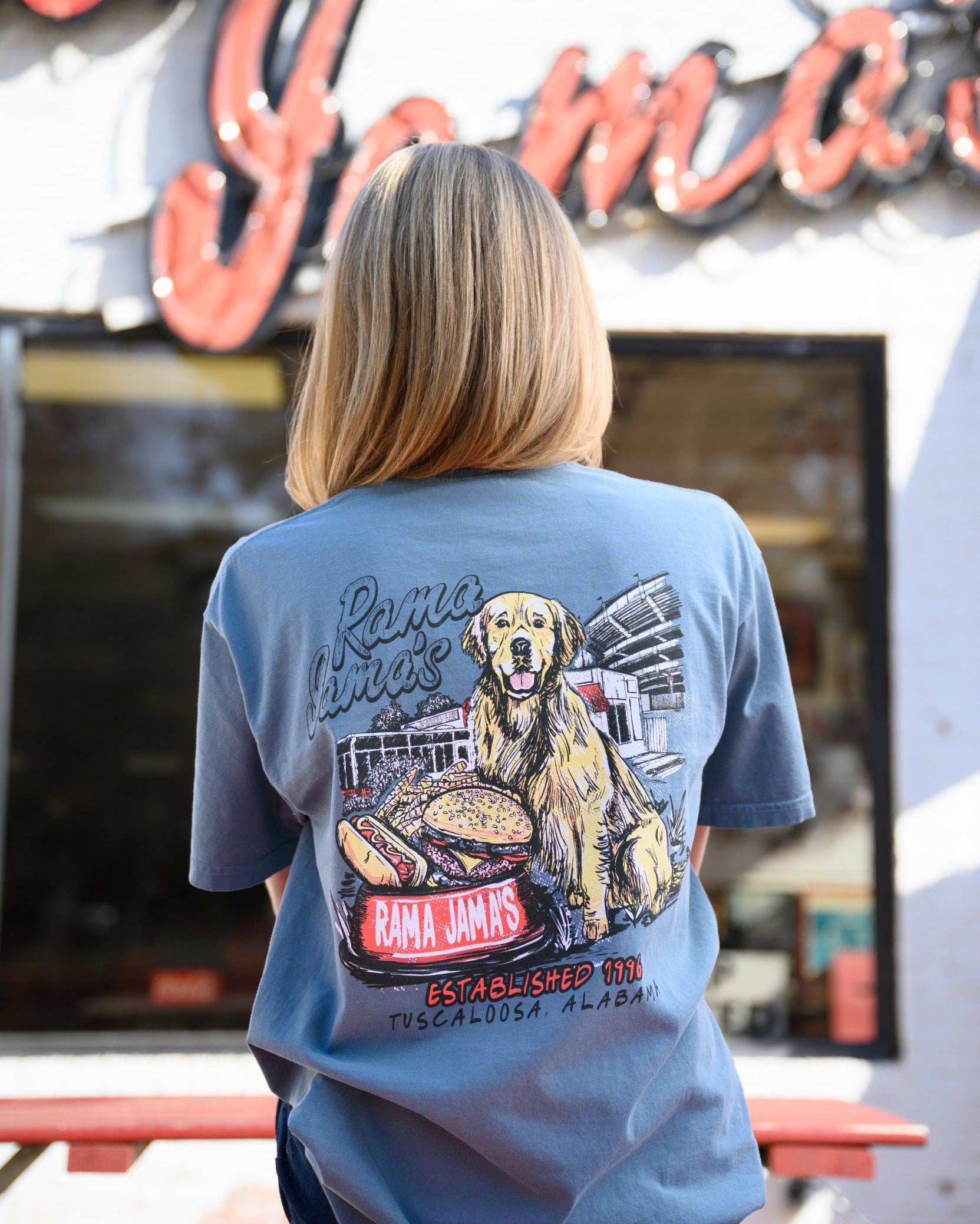 Rama Jama’s Dusty Blue shirt with an illustration of a golden retriever sitting at the picnic tables outside of the building with a cheeseburger and a hotdog