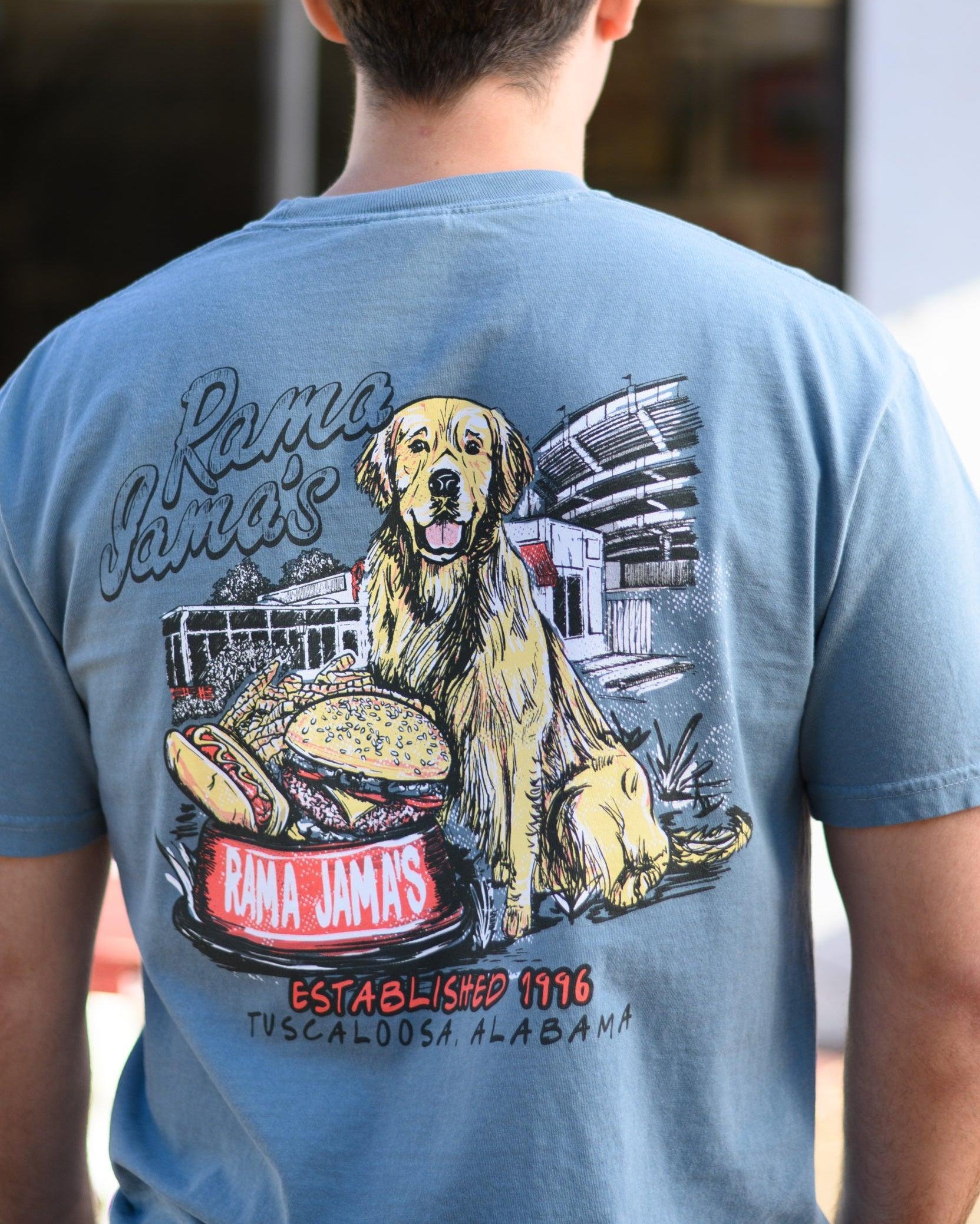Rama Jama’s Dusty Blue shirt with an illustration of a golden retriever sitting at the picnic tables outside of the building with a cheeseburger and a hotdog