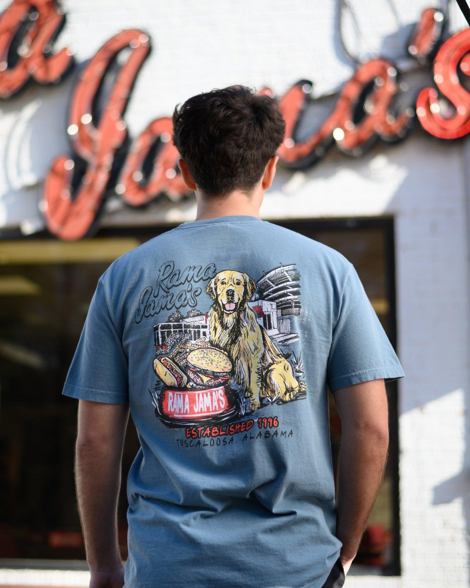 Rama Jama’s Dusty Blue shirt with an illustration of a golden retriever sitting at the picnic tables outside of the building with a cheeseburger and a hotdog