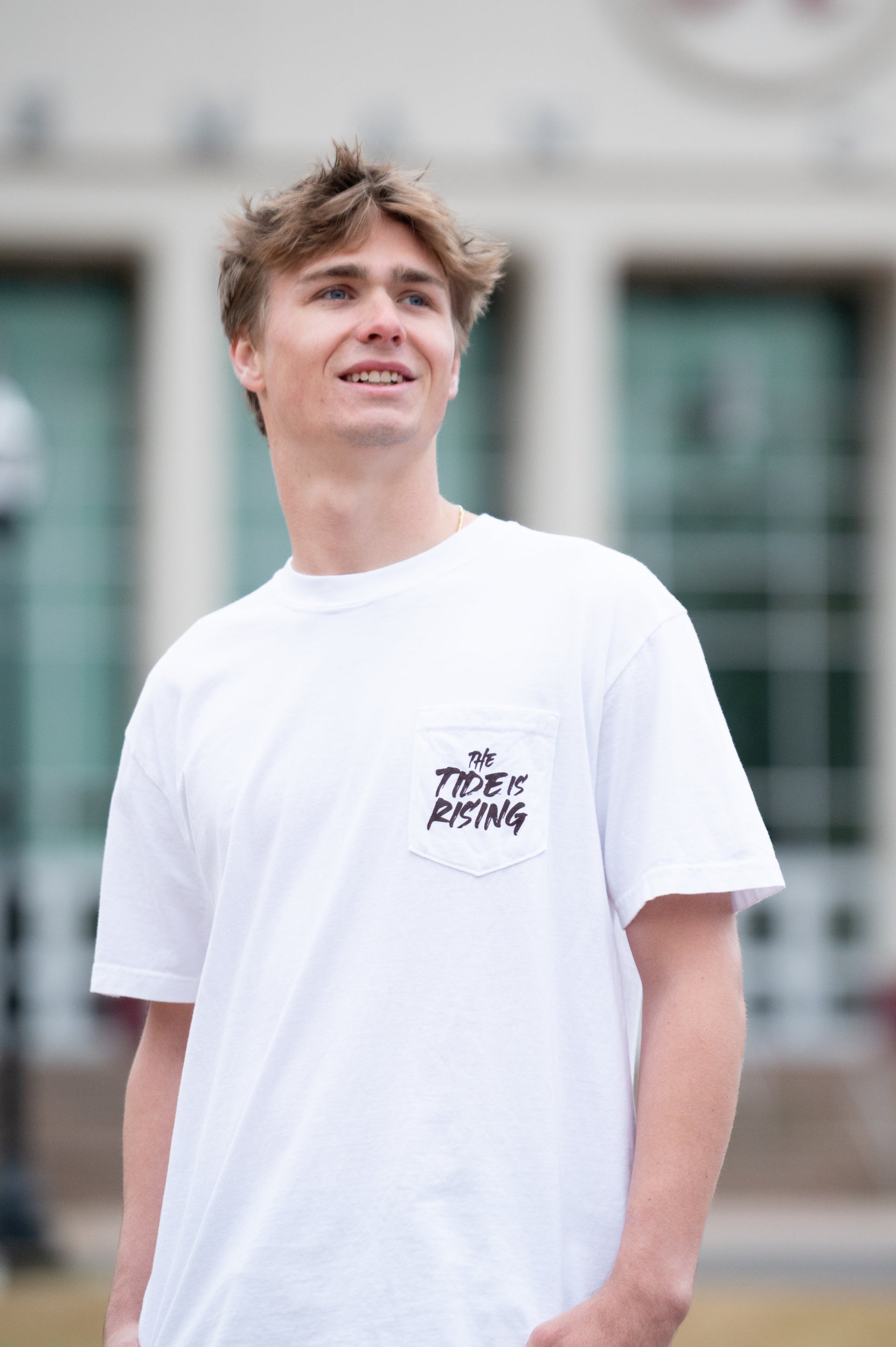 University of Alabama White T-shirt with a Script A brand folding chair that has a red flannel hanging over the back and a play planning board leaning against it. Below, Black text reading “The Tide is Rising”