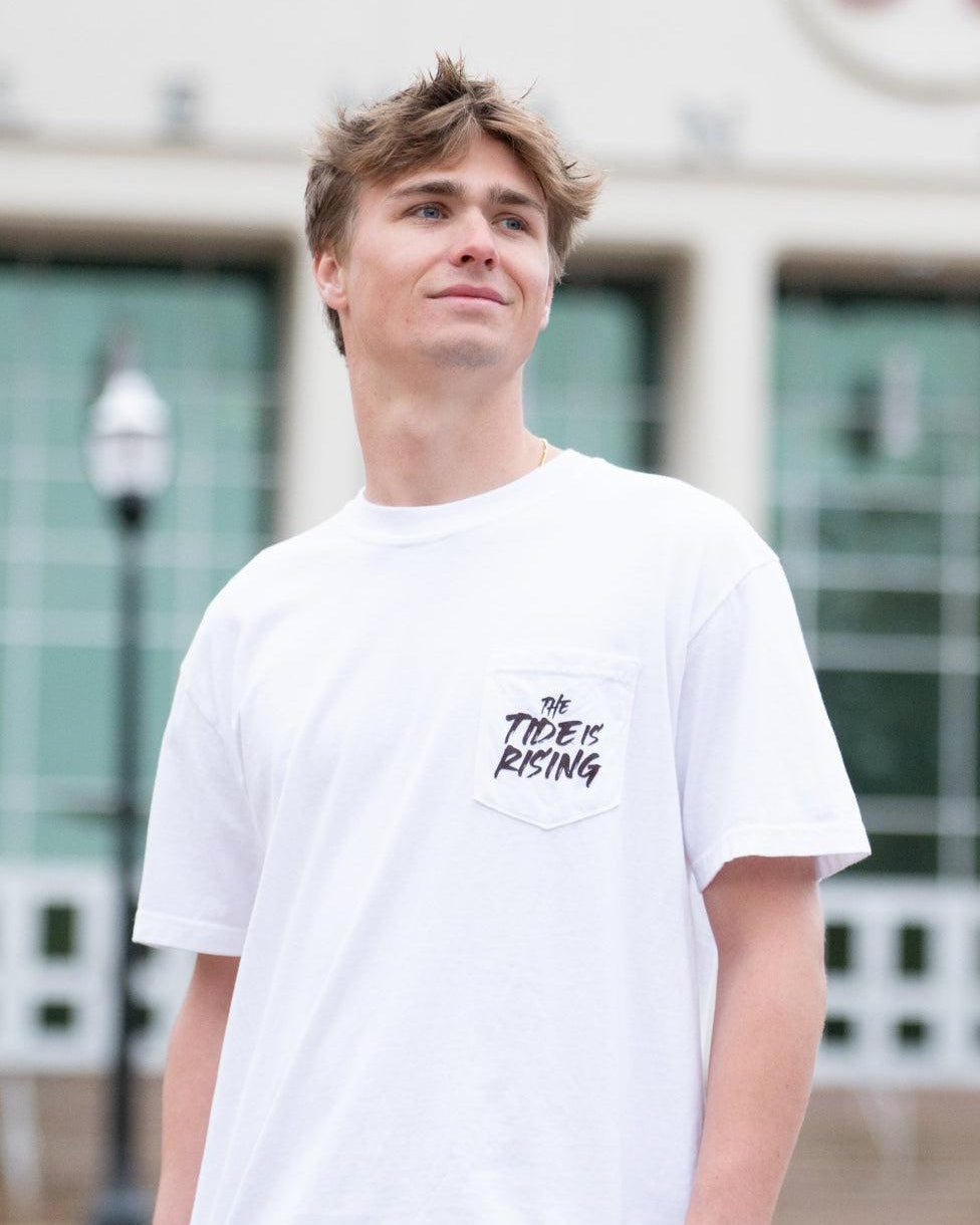 University of Alabama White T-shirt with a Script A brand folding chair that has a red flannel hanging over the back and a play planning board leaning against it. Below, Black text reading “The Tide is Rising”