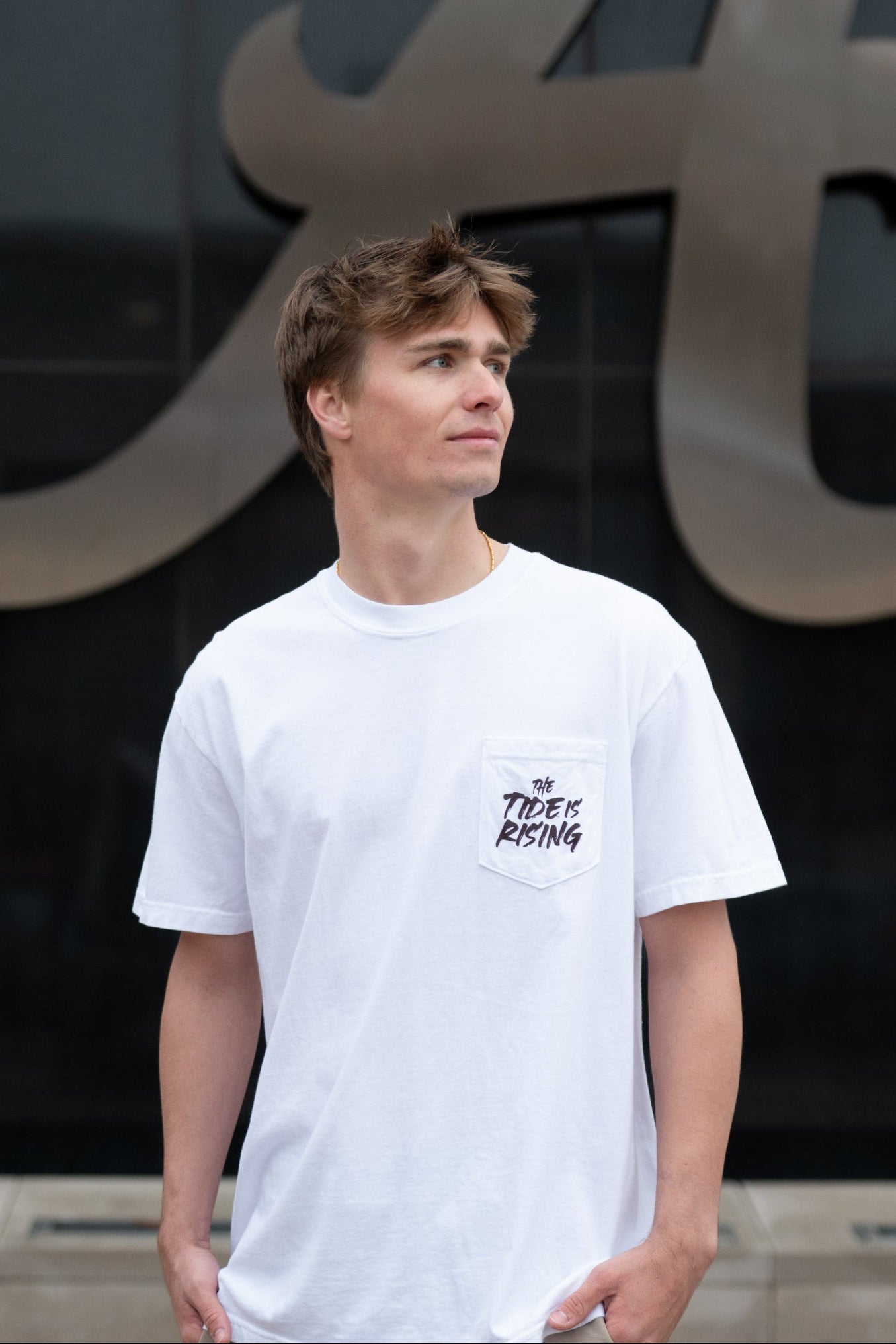 University of Alabama White T-shirt with a Script A brand folding chair that has a red flannel hanging over the back and a play planning board leaning against it. Below, Black text reading “The Tide is Rising”