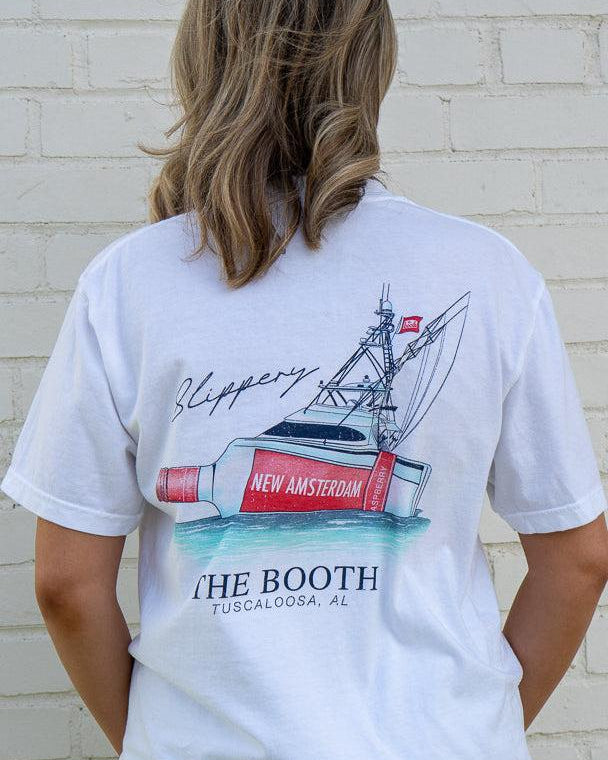 Person wearing a white t-shirt with a graphic design of a boat and text, standing against a light brick wall.