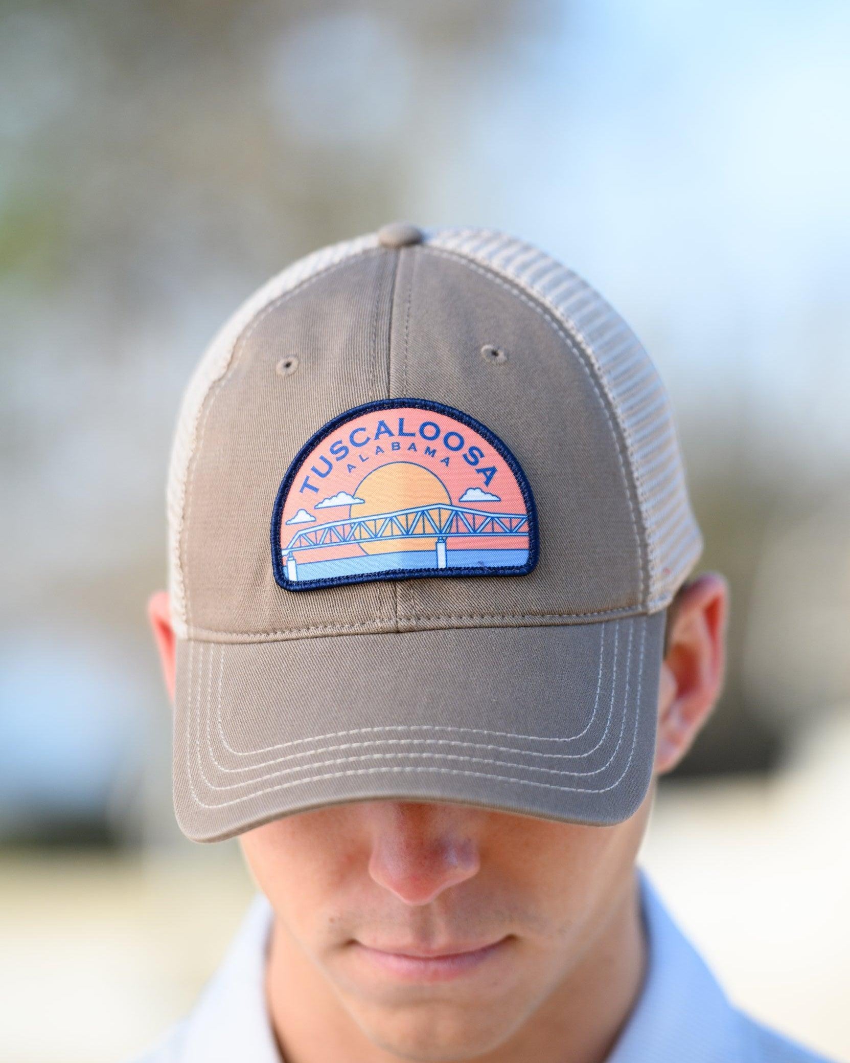 Tuscaloosa Beige Trucker Hat with a patch featuring Navy text "Tuscaloosa Alabama" a drawing of the sun setting on the iconic Tuscaloosa Trestle