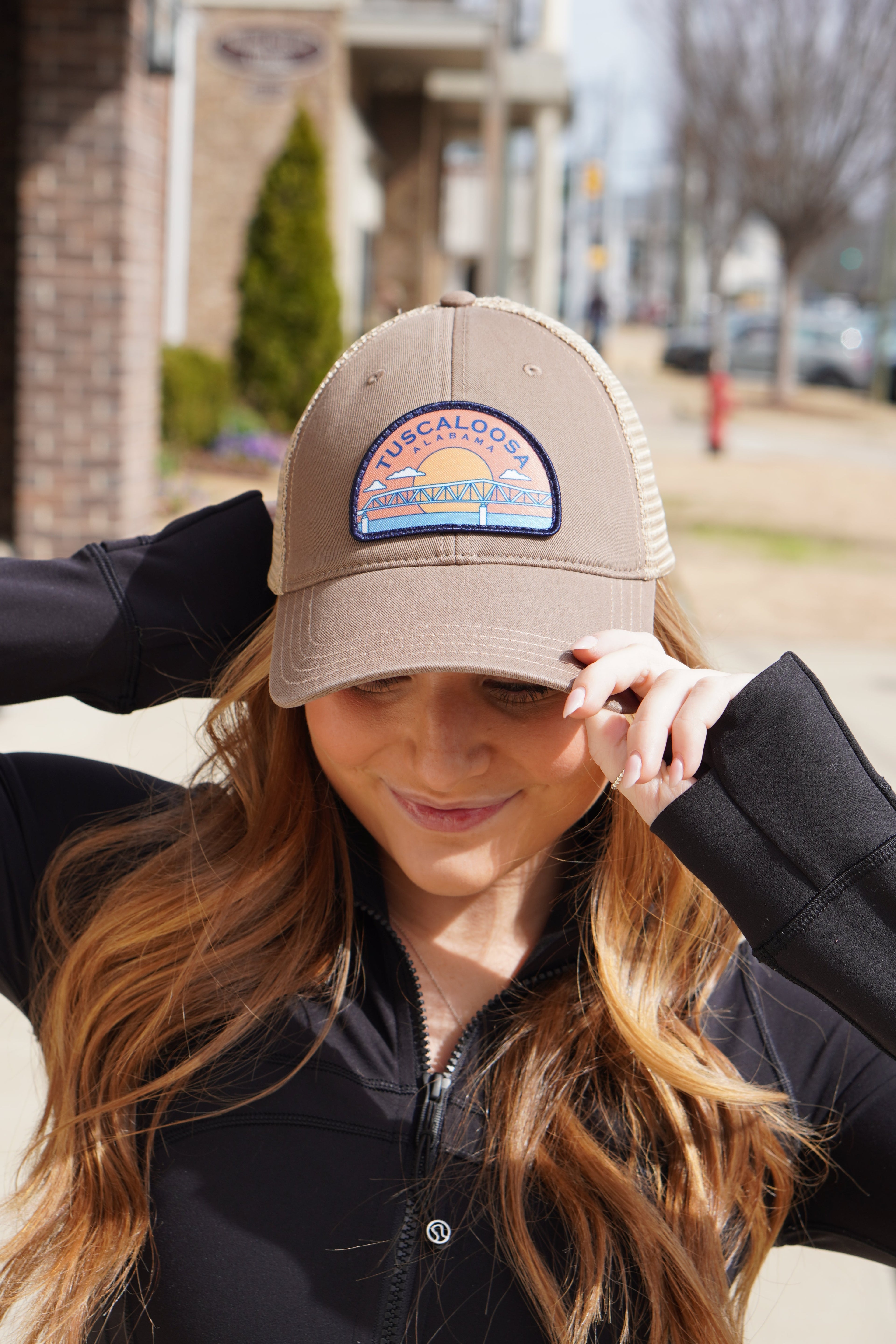 Tuscaloosa Beige Trucker Hat with a patch featuring Blue text "Tuscaloosa Alabama" a drawing of the sun setting on the iconic Tuscaloosa Trestle