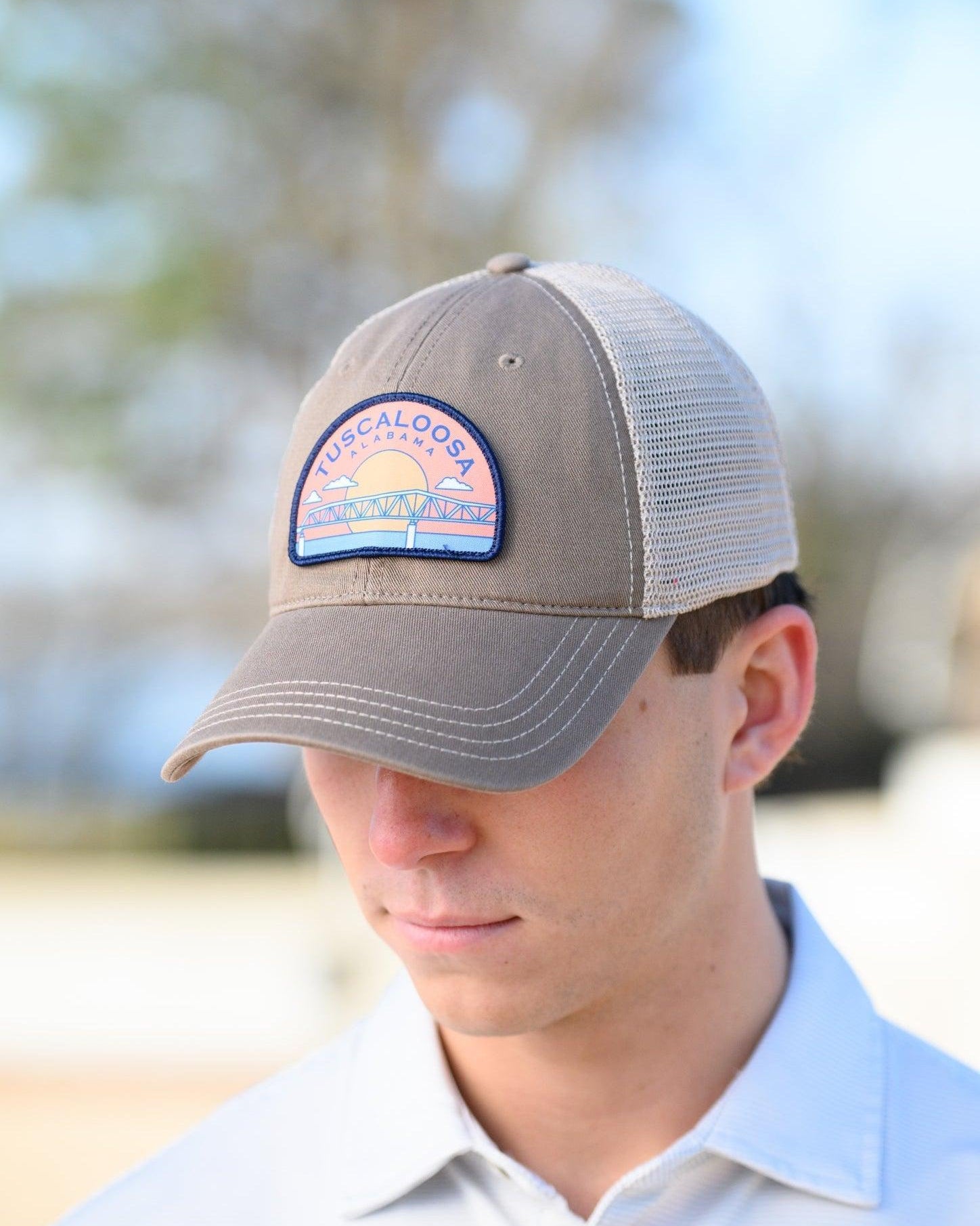 Tuscaloosa Beige Trucker Hat with a patch featuring Navy text "Tuscaloosa Alabama" a drawing of the sun setting on the iconic Tuscaloosa Trestle