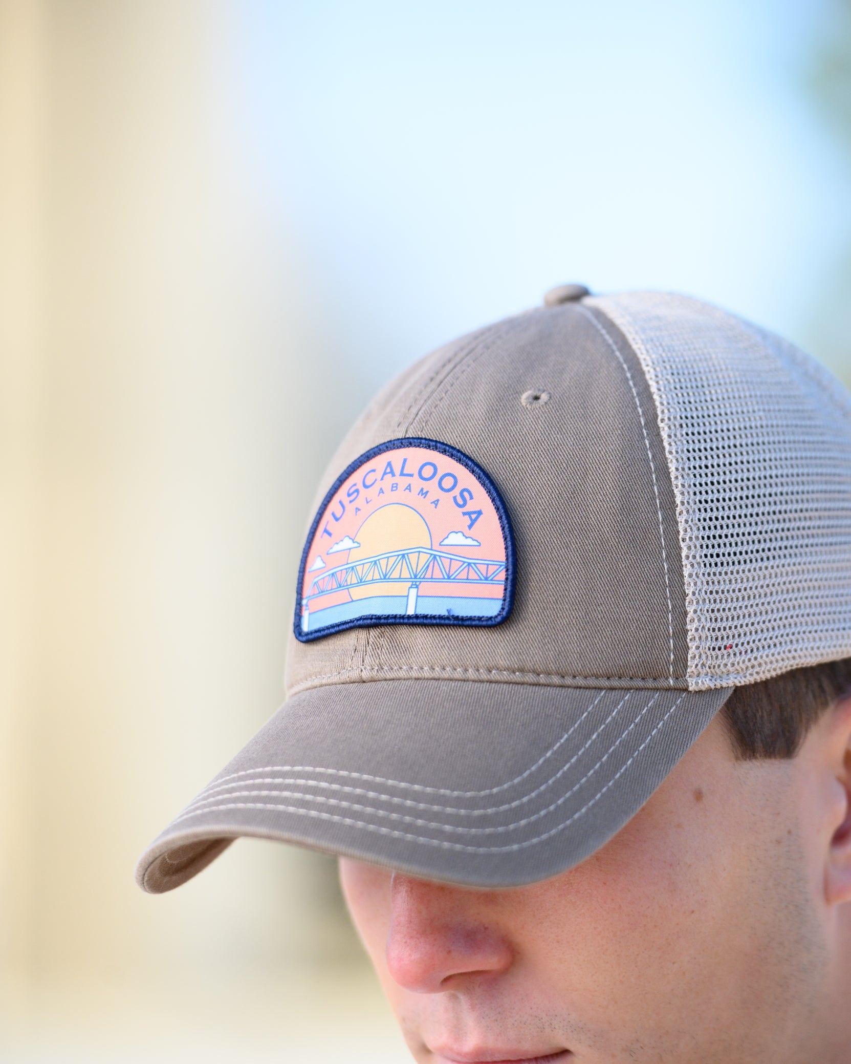 Tuscaloosa Beige Trucker Hat with a patch featuring Navy text "Tuscaloosa Alabama" a drawing of the sun setting on the iconic Tuscaloosa Trestle