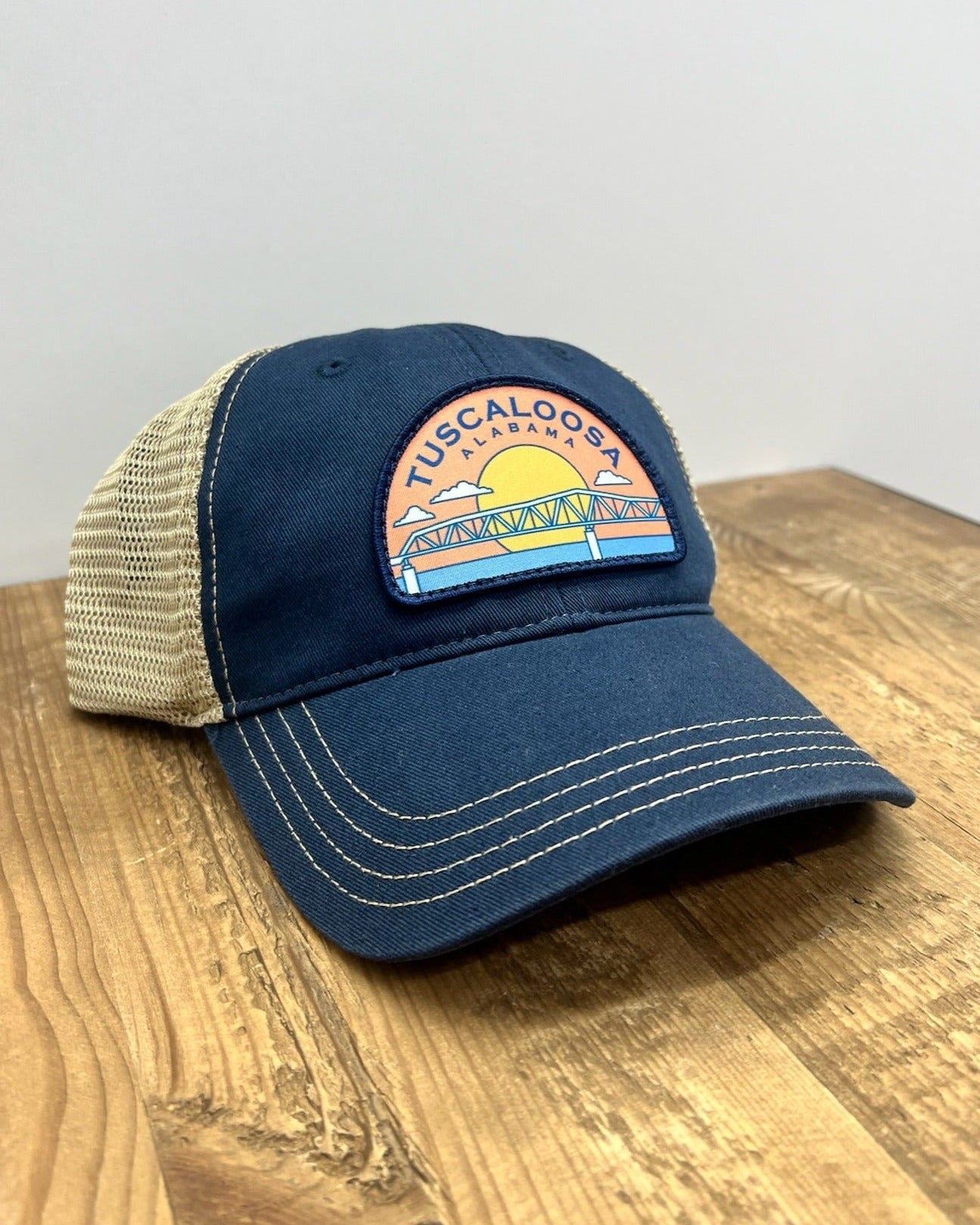 Tuscaloosa Navy and Beige Trucker Hat with a patch featuring Blue text "Tuscaloosa Alabama" a drawing of the sun setting on the iconic Tuscaloosa Railroad Tracks