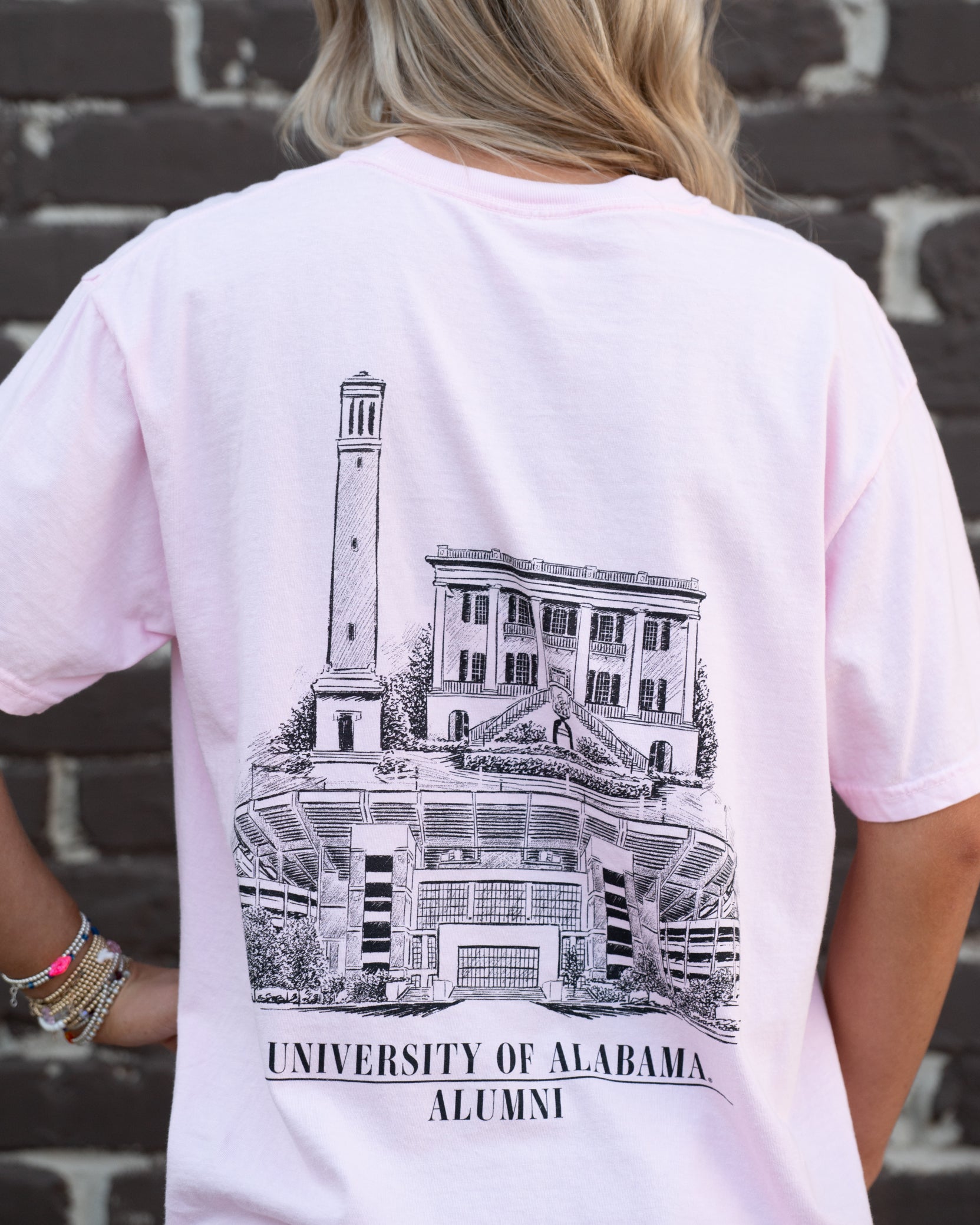 University of Alabama Alabama Alumni pink Comfort Colors T-Shirt with a graphic of Bryant-Denny Stadium, the President's Mansion, and Denny Chimes, and the text 'UNIVERSITY OF ALABAMA ALUMNI'.