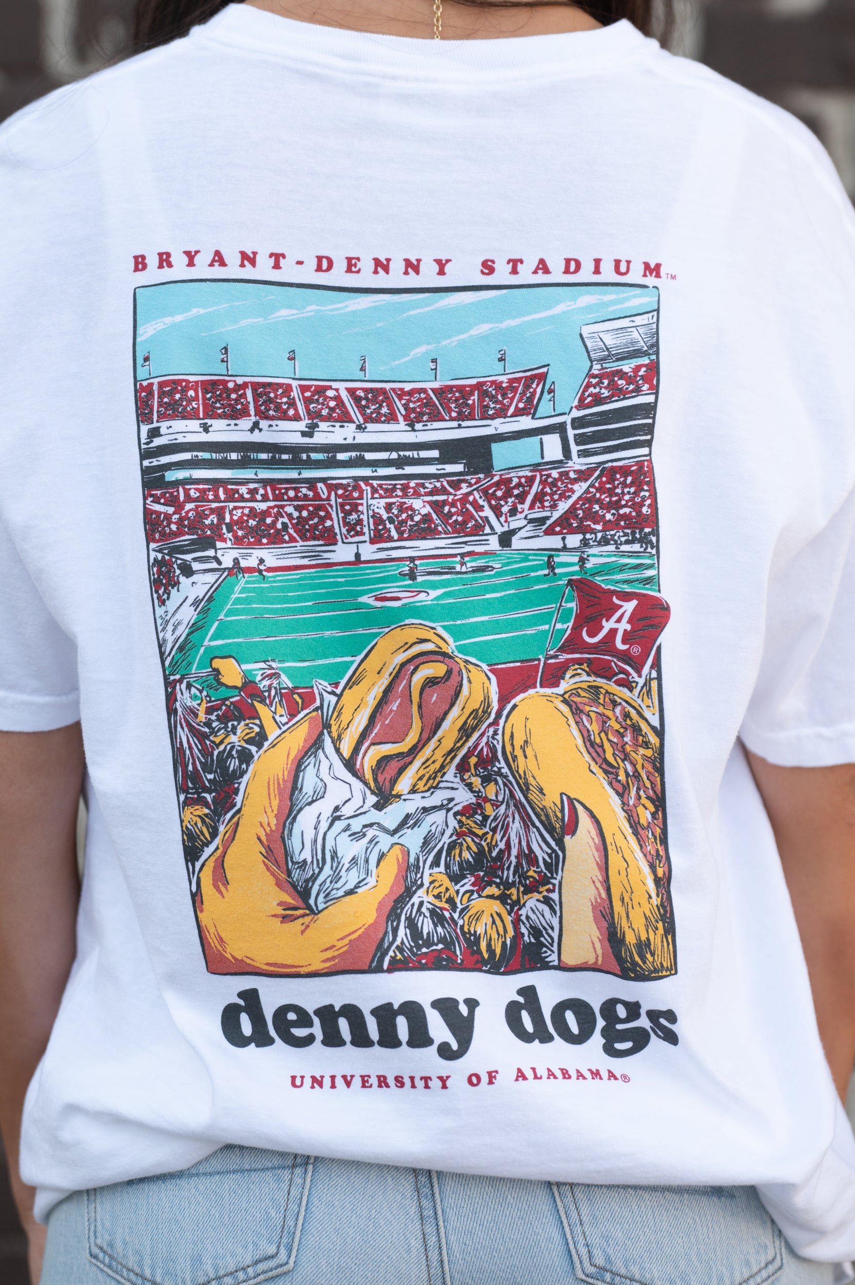 University of Alabama White Denny Dogs Comfort Colors T-Shirt with an image of two hands holding hot dogs at Bryant- Denny Stadium above the Black text "denny dogs"