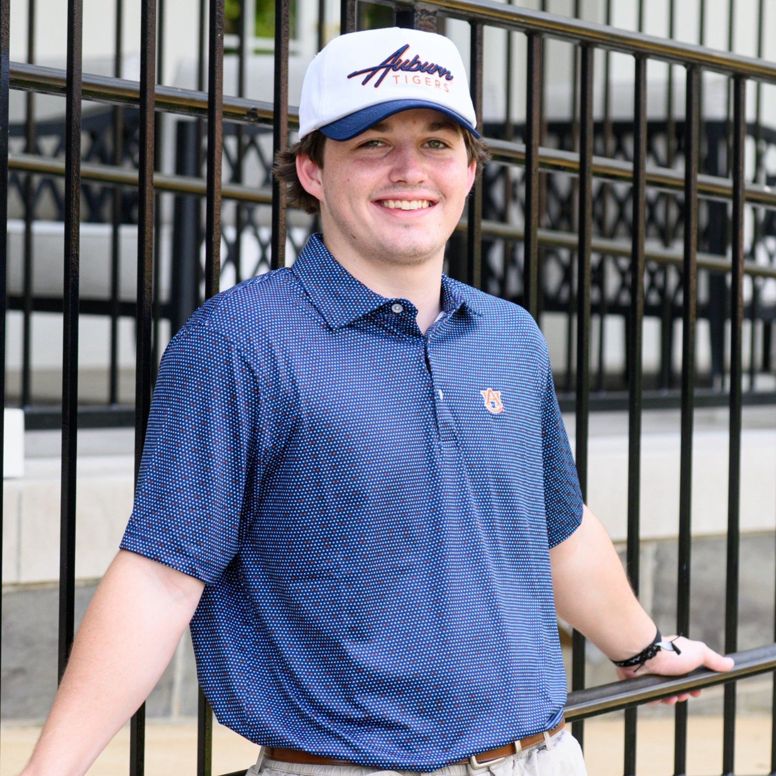 Man leaning against metal railing wearing white and blue hat with Auburn script across the front and a blue diamond patterned polo shirt with Auburn University's block AU logo on the left chest.