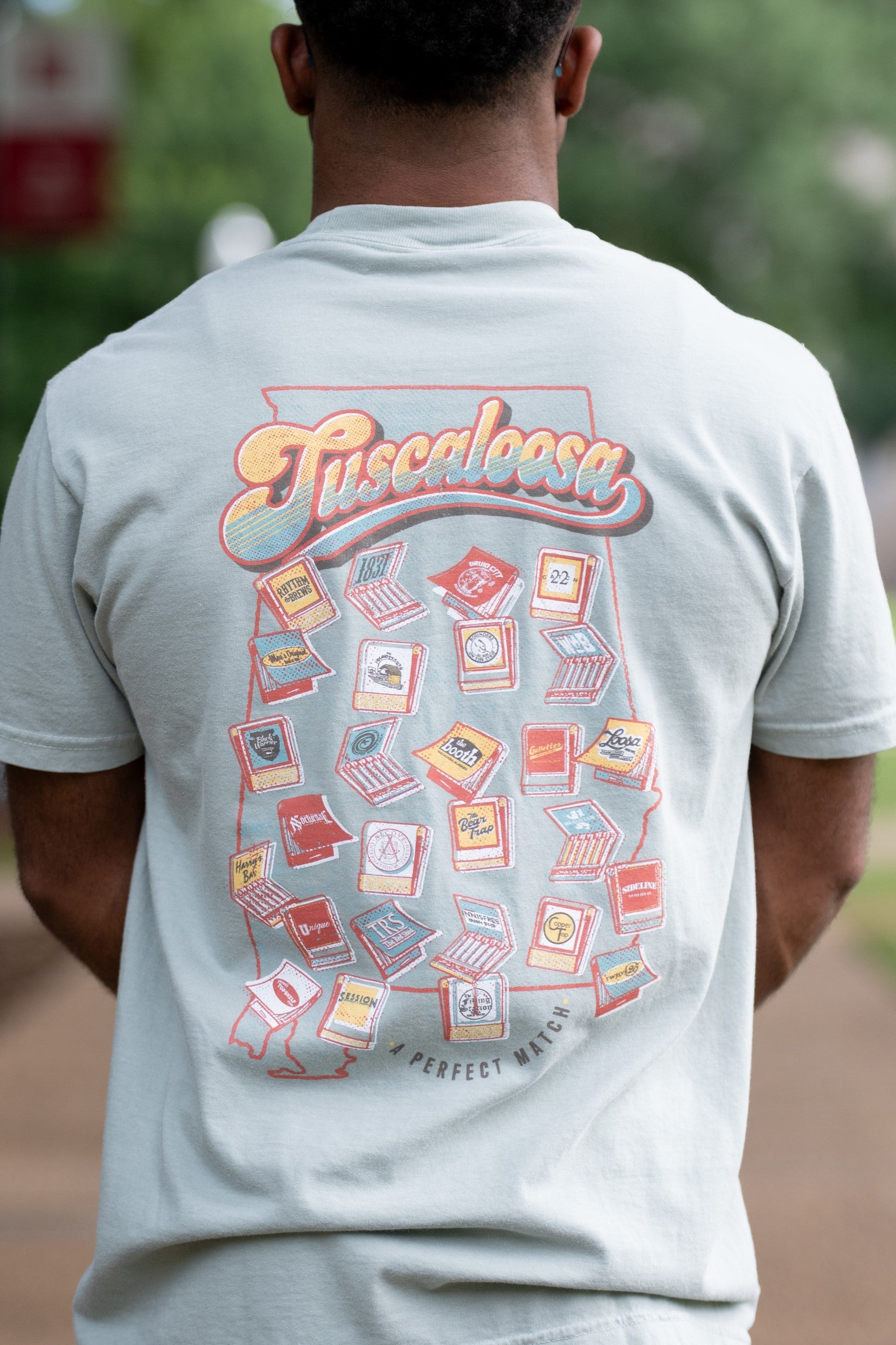 A Perfect Match Green Comfort Colors T-Shirt  with the text "Tuscaloosa" and images of matchbooks inside of an outline of the state of Alabama and smaller text reading "A PERFECT MATCH" outside the outline 