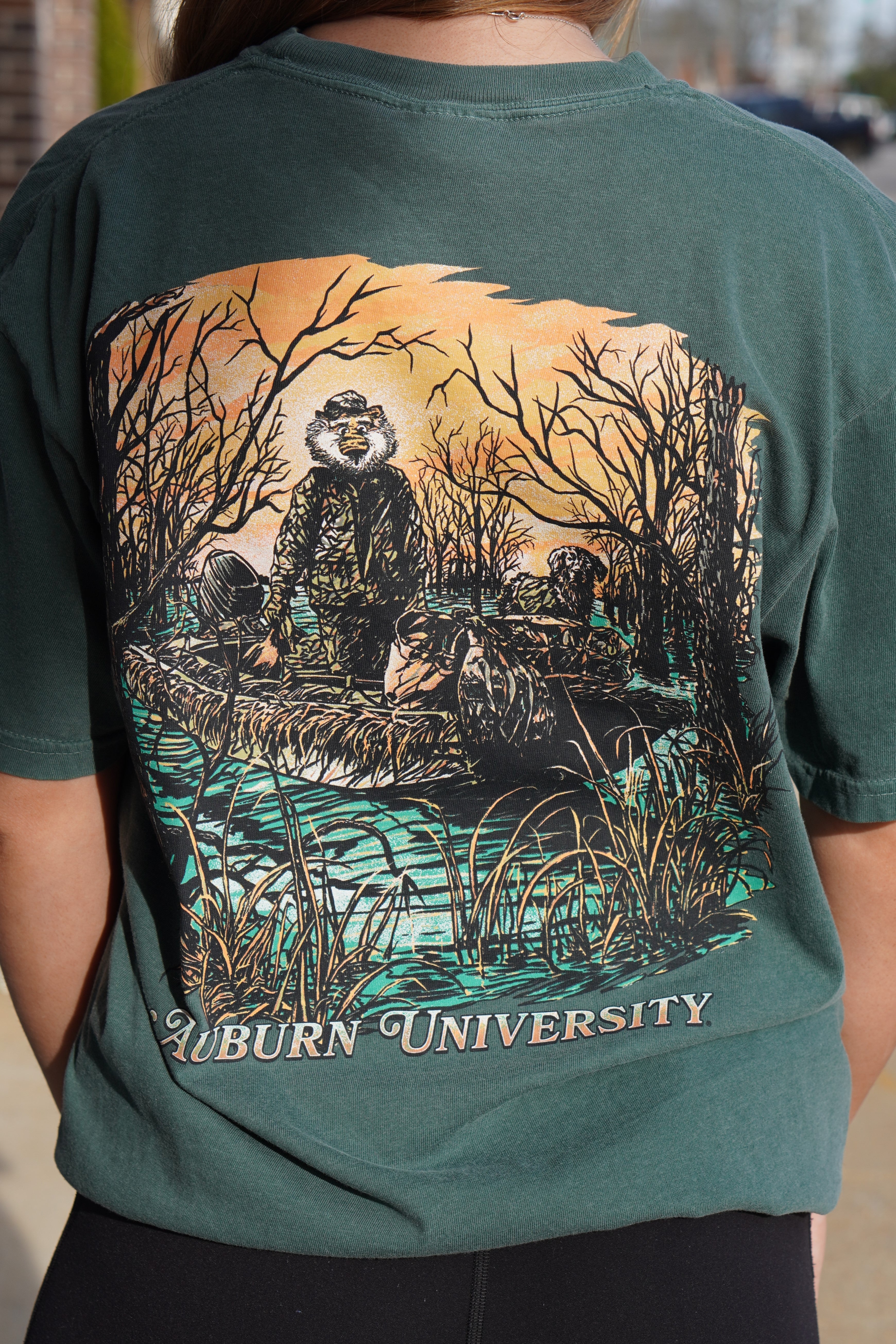 Auburn University Aubie Duck Hunt Blue Spruce Comfort Colors T-Shirt with an image of Aubie the Tiger wearing hunting gear with a dog in a boat in the swamp 