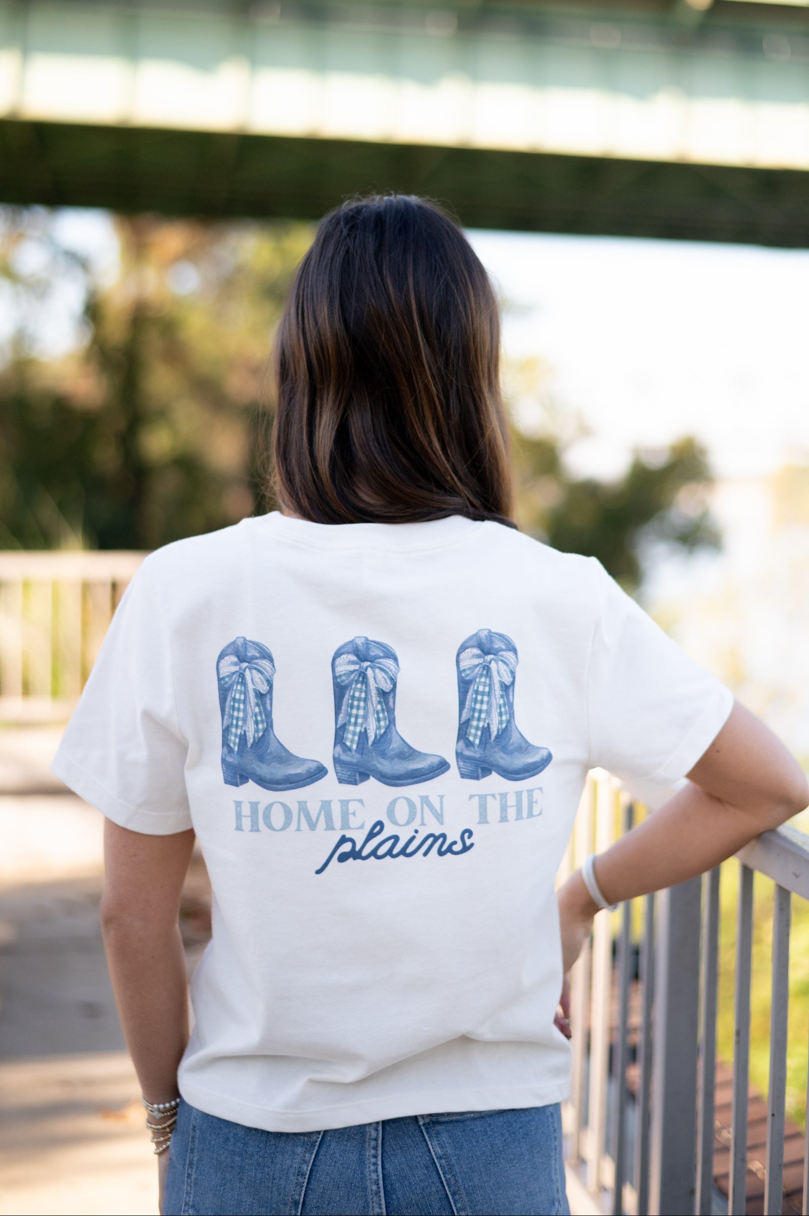 Auburn University White Ladies Tee with a vintage Blue illustration of cowgirl boots with bows tied on them above Blue text “Home On The Plains”