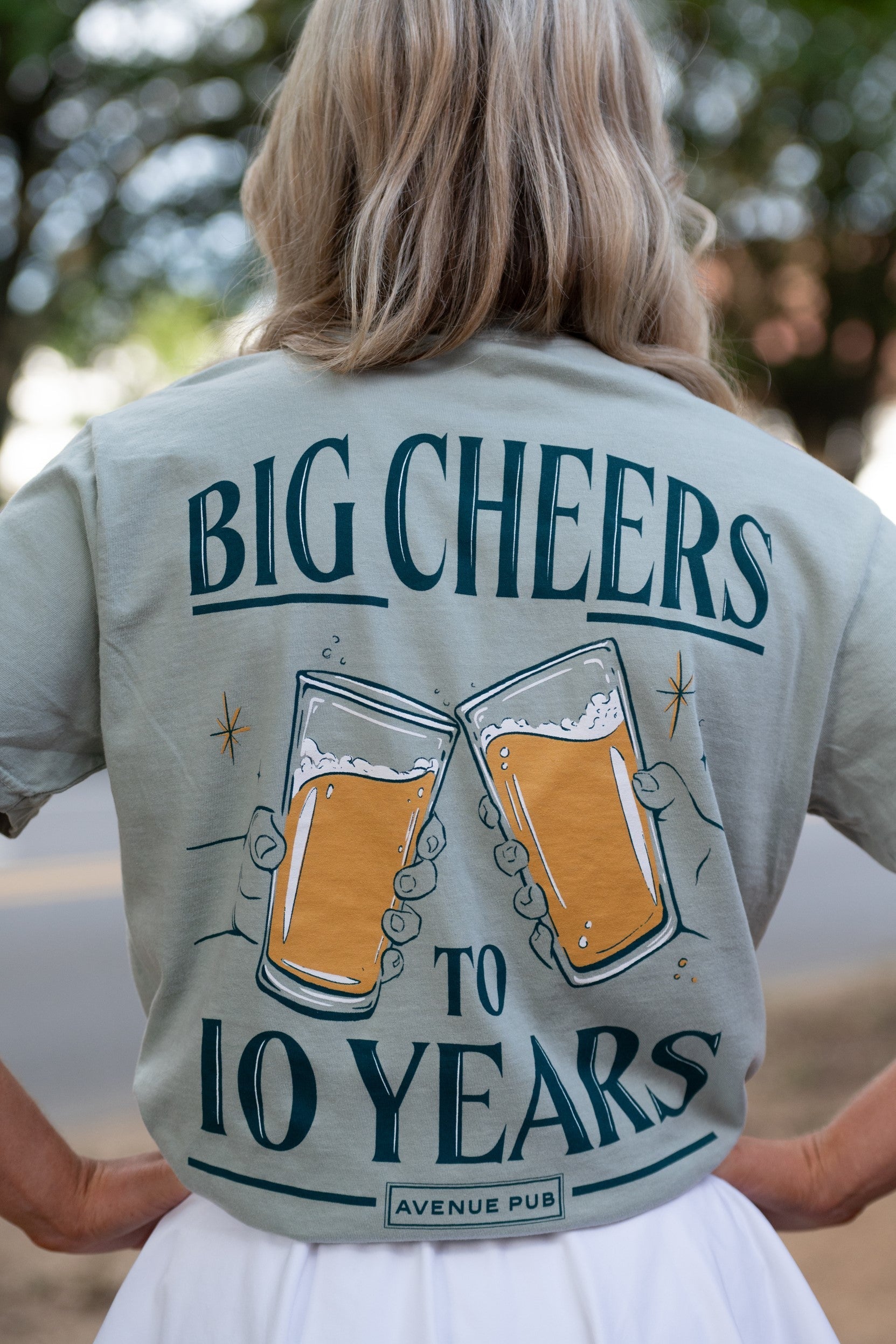 Avenue Pub's Beer Cheers Light Green Comfort Colors T-Shirt with an image of  people clinking glasses of beer in between text "Big Cheers to 10 Years" (Back) 