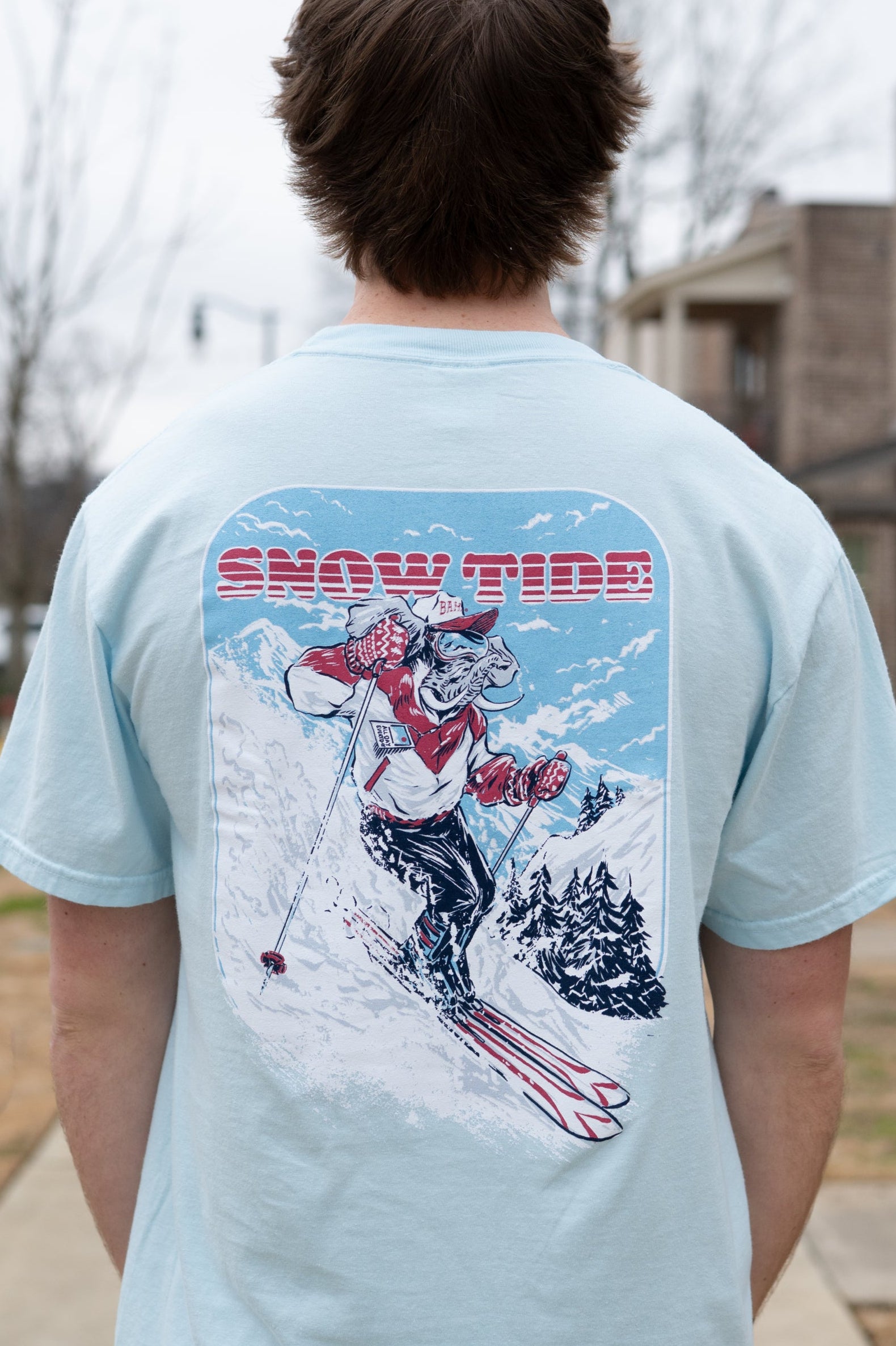 University of Alabama Light Blue T-Shirt with text "Snow Tide" above an image of Big Al in a ski outfit skiing down a snow-covered mountain