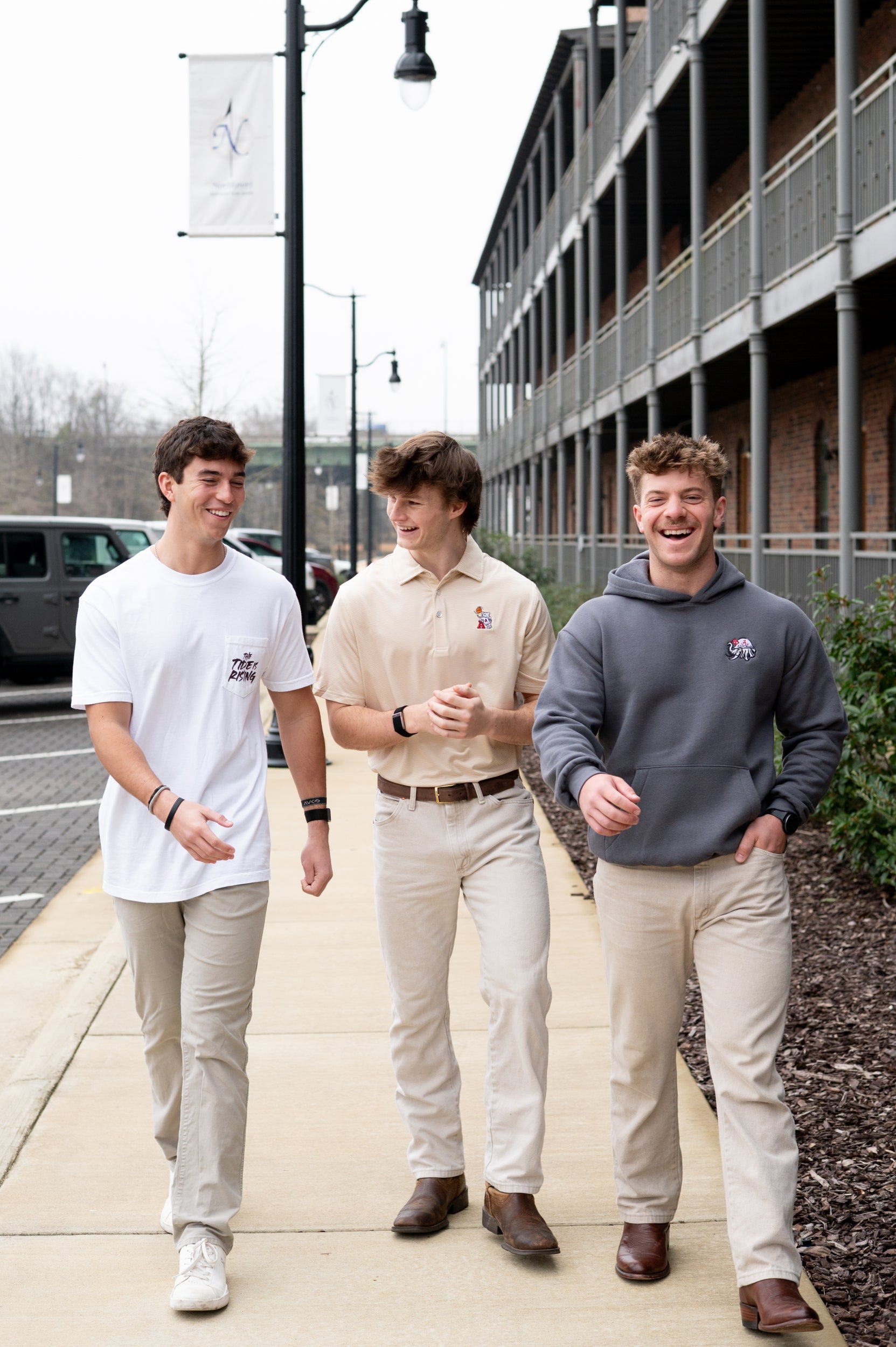 Three young men walking together on a sidewalk, with one wearing a white shirt, another in a beige Polo, and the third in a gray hoodie.