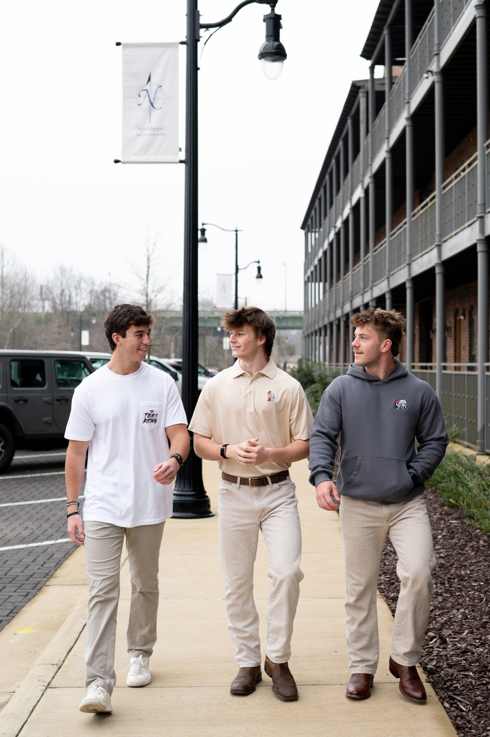 Three young men walking together on a sidewalk, with one wearing a white shirt, another in a beige Polo, and the third in a gray hoodie.