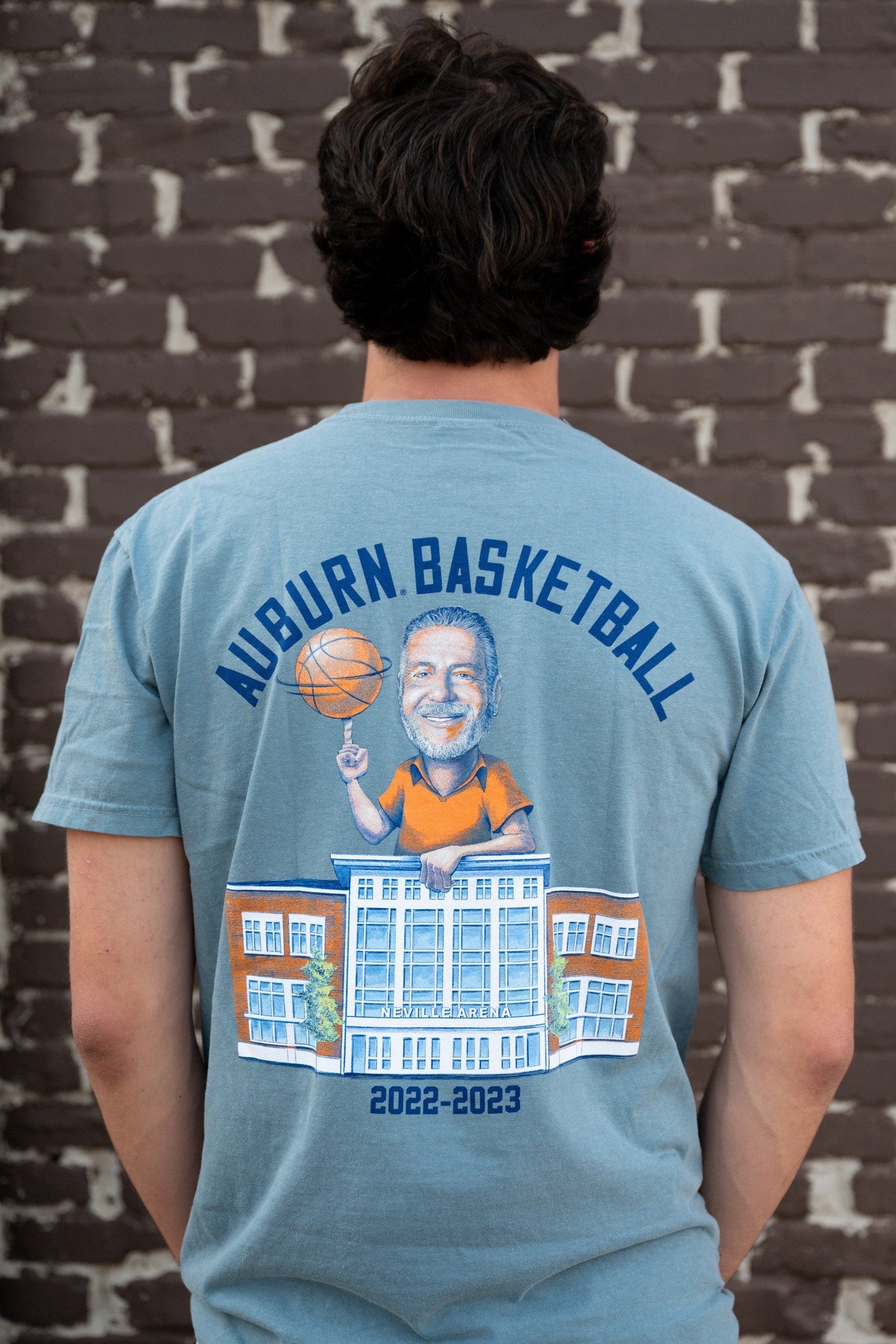 Auburn University Blue Bruce Pearl 2023 Comfort Colors T-Shirt with Navy text "Auburn Basketball" above an image of Auburn Basketball Coach Bruce Pearl spinning a basketball and leaning on the basketball building 