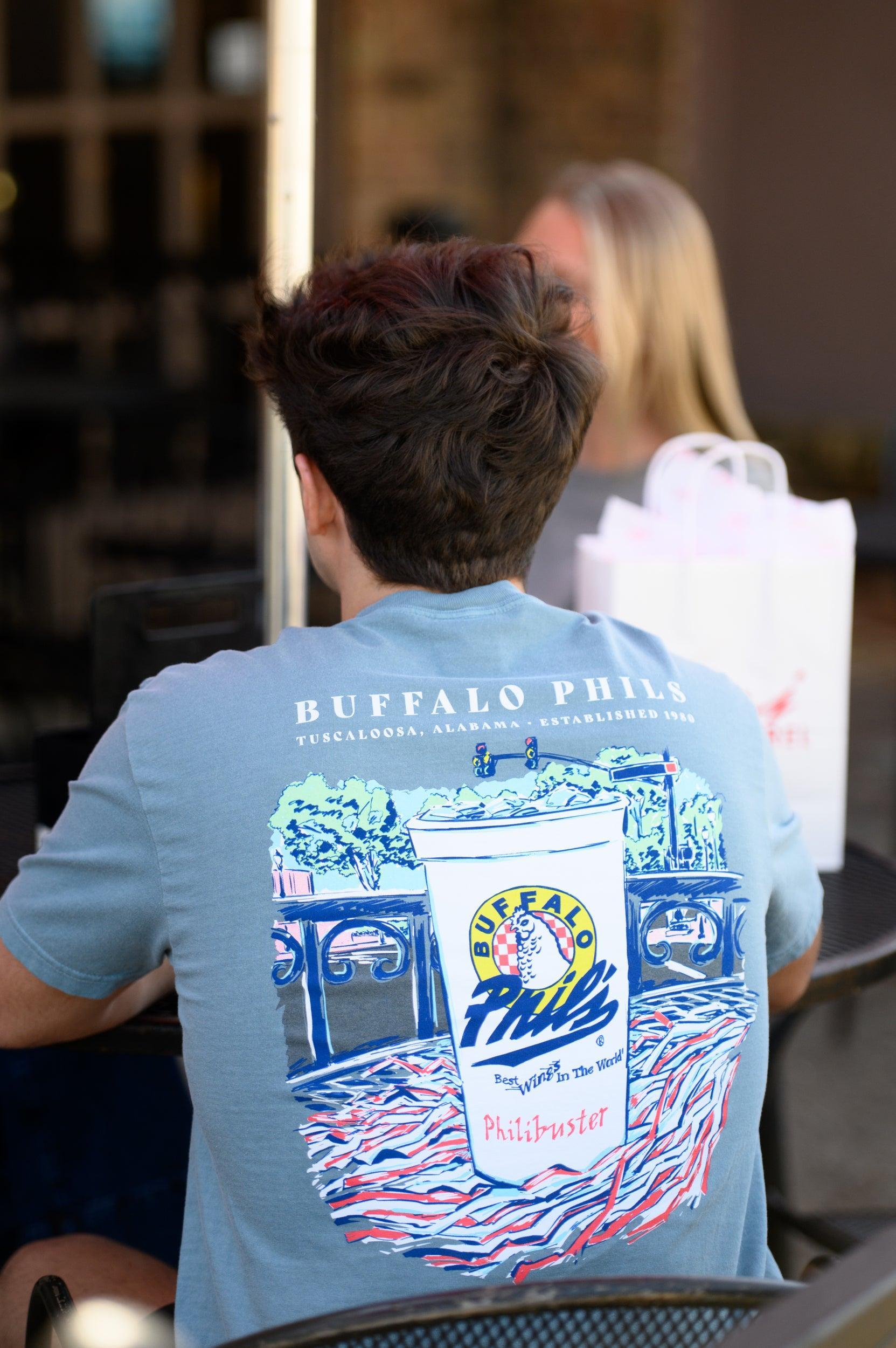 Ice Blue Buffalo Phil's T-Shirt with an image of a Buffalo Phil's cup sitting on top of a shaker on a table looking out on The Strip
