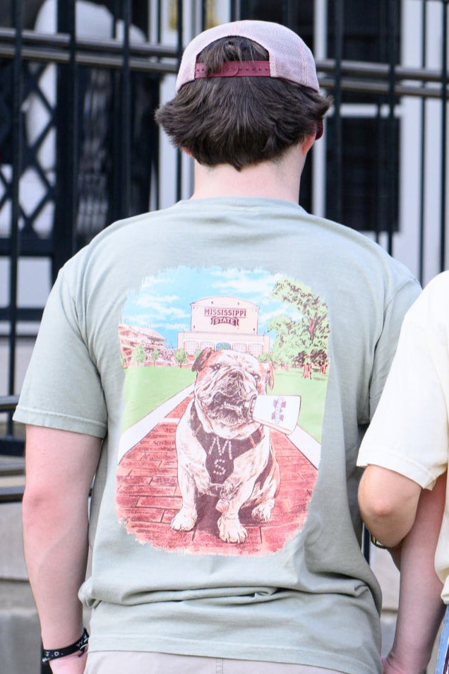Mississippi State Tan T-Shirt with an image of Bully the bulldog holding a cowbell in it’s mouth on the brick walk into campus