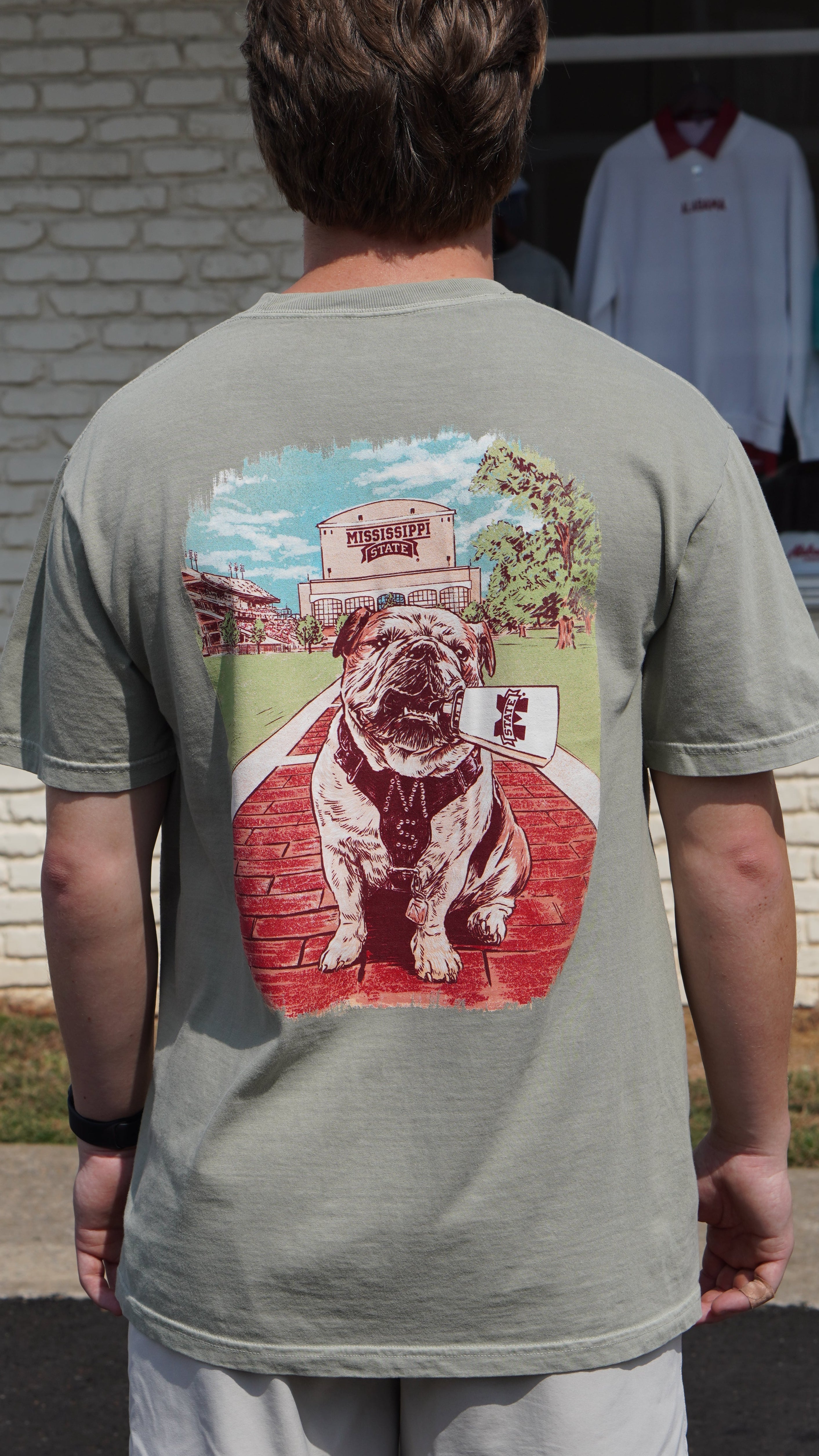 Mississippi State Tan T-Shirt with an image of Bully the bulldog holding a cowbell in it’s mouth on the brick walk into campus