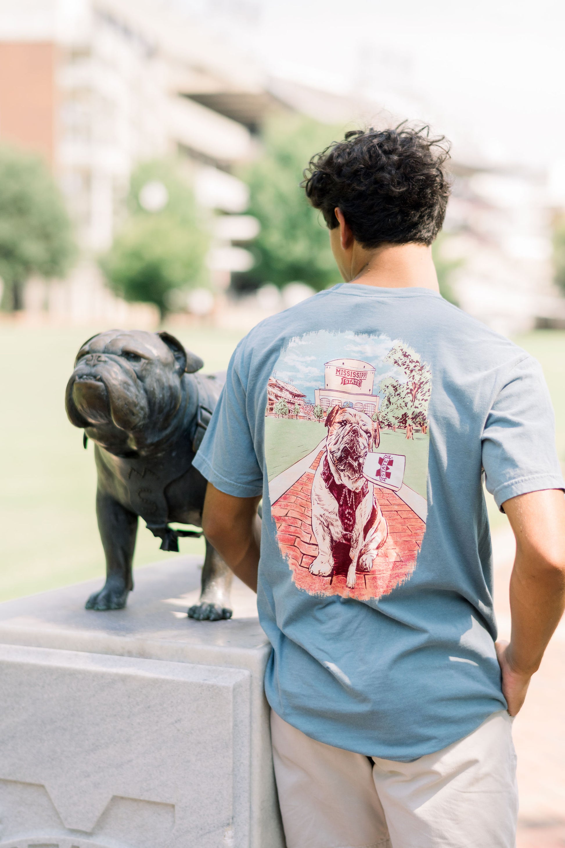Mississippi State Dusty Blue T-Shirt with an image of Bully the bulldog holding a cowbell in its mouth on the brick walk into campus